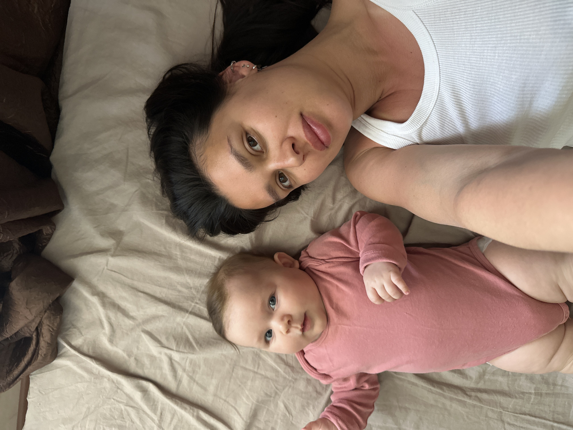 A fair-skinned woman and a baby are lying on a bed. The woman, in a white tank top, is on the upper part of the image. Her dark hair is spread out, and she is looking at the camera. The baby, wearing a pink long-sleeved onesie, is below her. The bed has light brown sheets and a dark brown blanket on the left. There is no indication that this image was taken in Chaikovsky, Russia.
