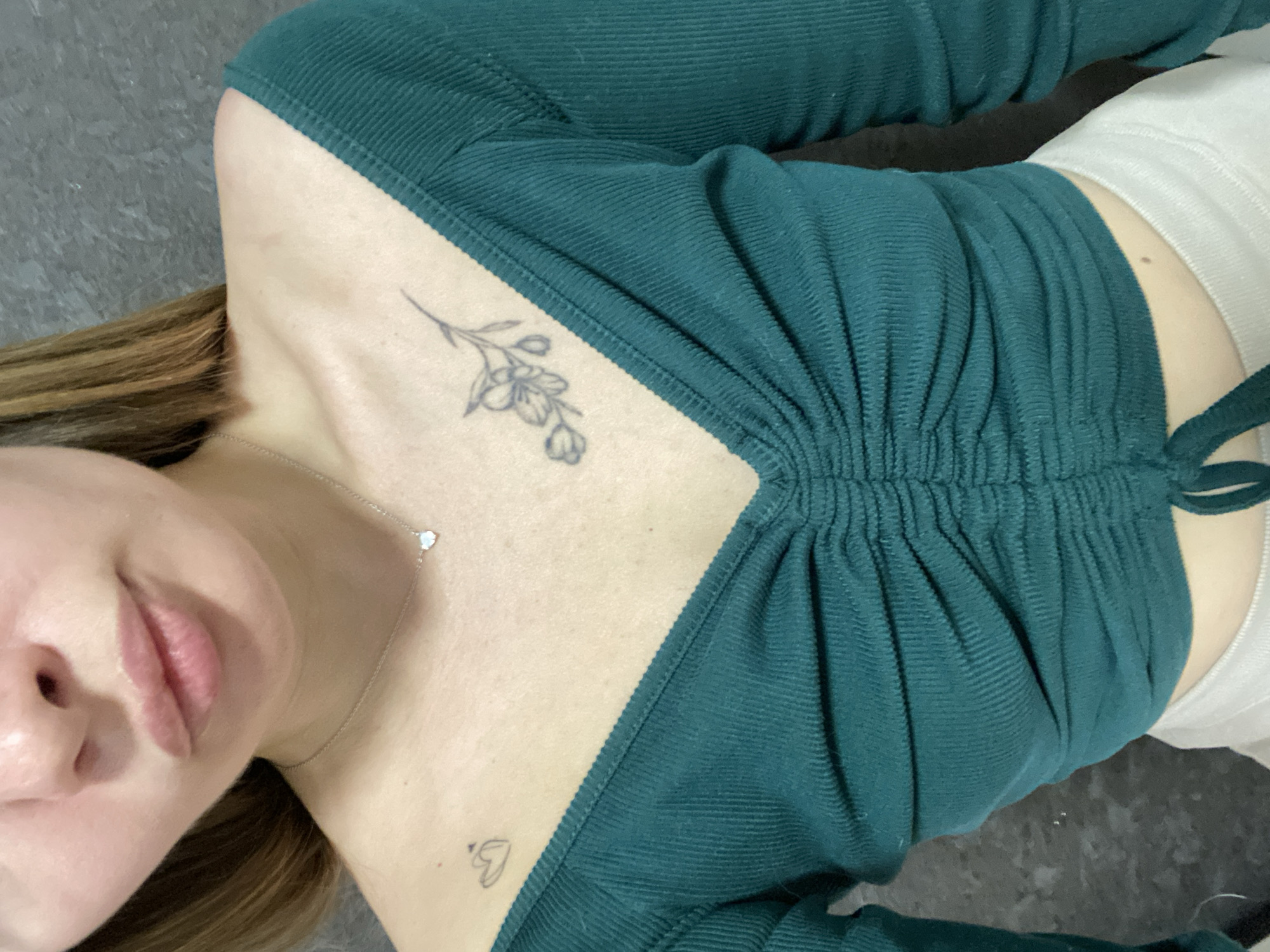 A woman with light brown hair is lying on a gray surface. She is wearing a teal, long-sleeved, ribbed shirt that is ruched in the center. She has a flower tattoo on her upper chest, a heart-shaped tattoo on her lower chest, and a silver necklace with a heart pendant. She is also wearing white shorts. The background is out of focus. There is no indication of a location in Izhevsk, Russia.