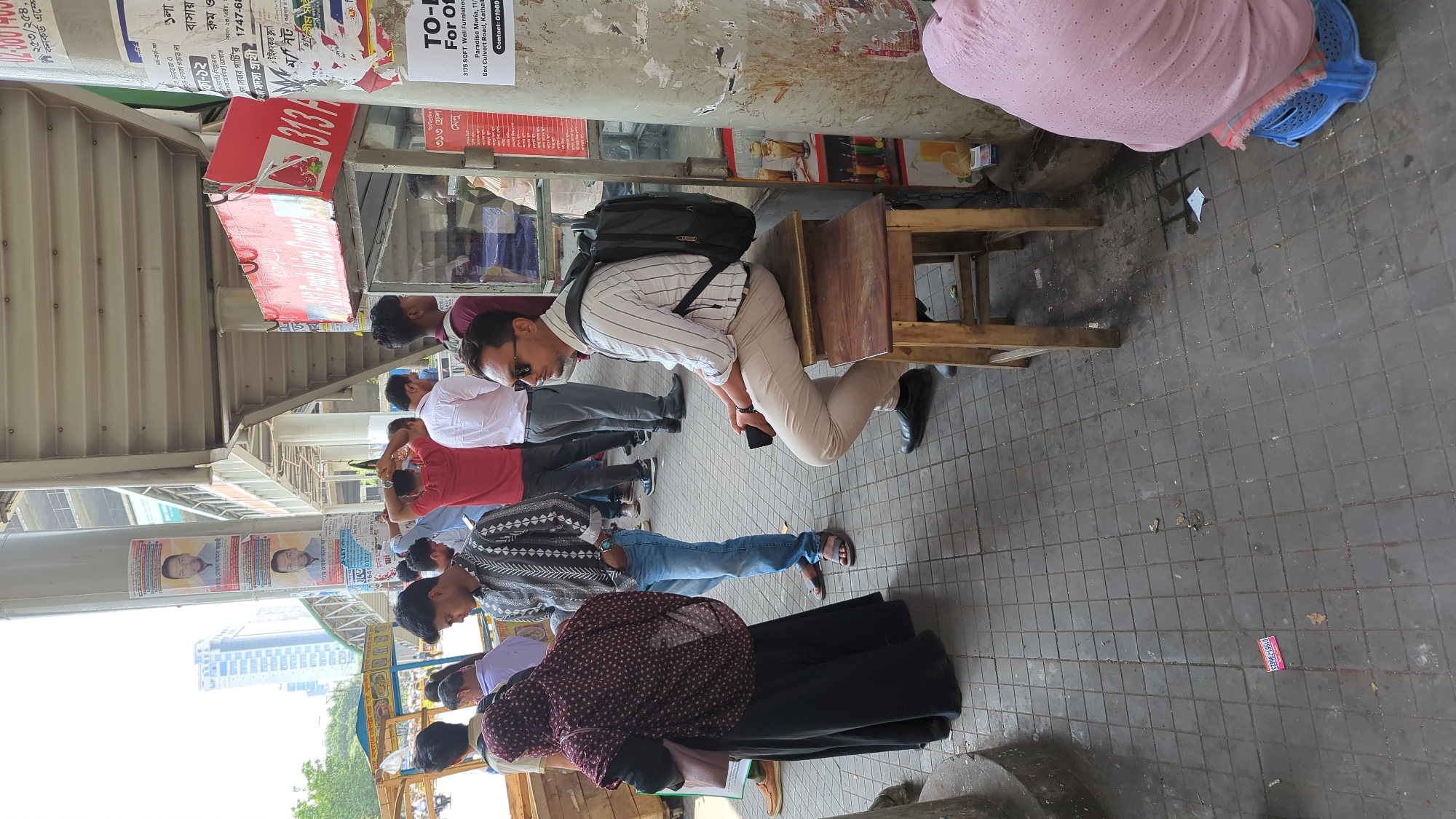 Here is a description of the image:
The image captures a street scene in Dhaka, Bangladesh. A line of individuals appears to be waiting, perhaps for a service or transportation. One man is seated on a wooden stool, wearing a striped shirt, tan pants, and black shoes. He has a backpack and is looking down while holding a smartphone. Other people stand nearby, dressed in various styles of clothing, including casual shirts and jeans. A woman is wearing a black skirt and a patterned top.
The scene takes place under a covered walkway with posters and advertisements attached to the pillars and walls. Buildings, including high-rise structures, can be seen in the background, indicating an urban area. A food stand with the number 313 on it is visible.