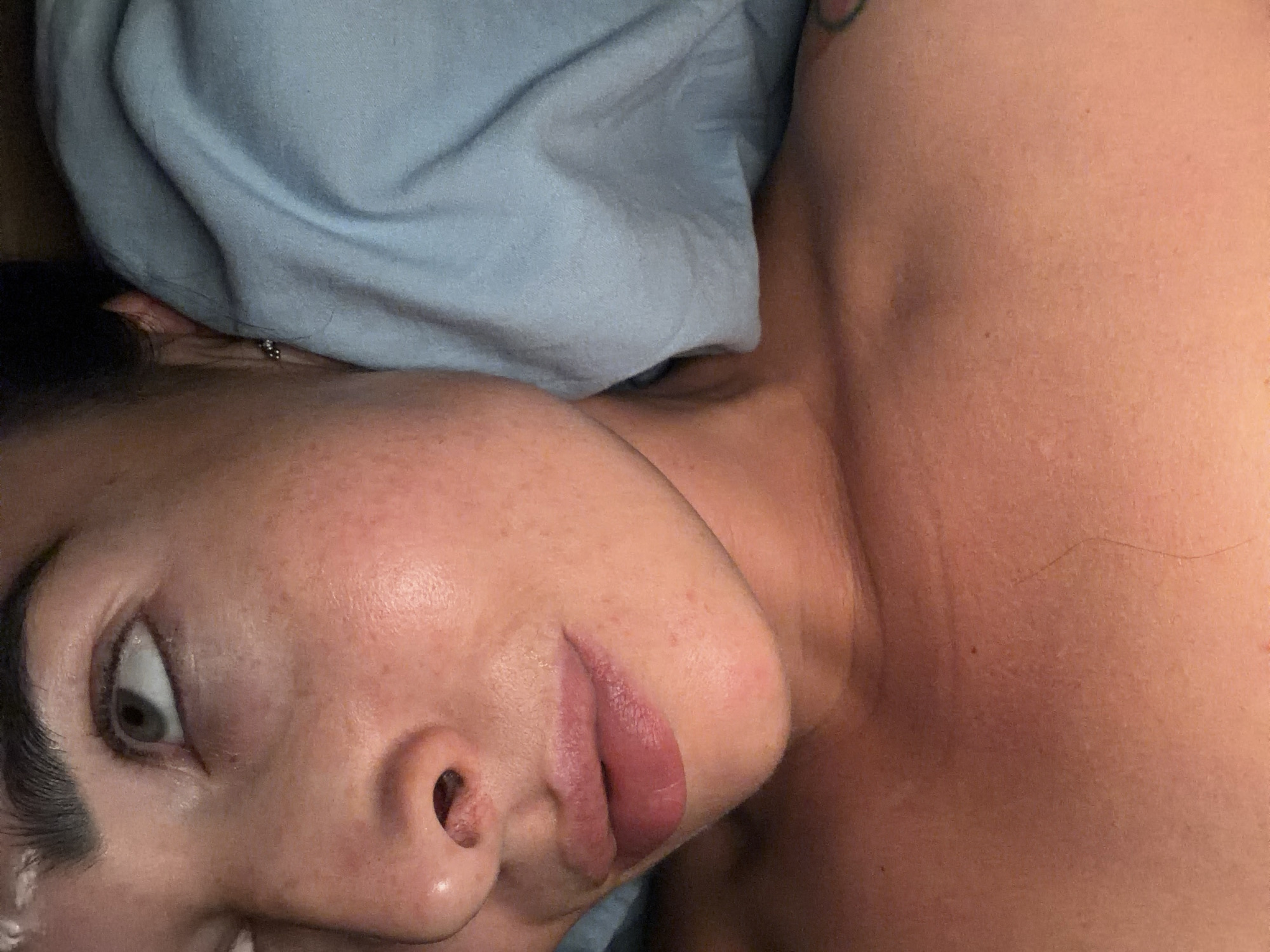 A close-up, angled photograph depicts a person lying on their side, with their head resting on a light blue pillow. The person's face is visible, showing an eye looking upwards, a nose, and lips. Their skin has freckles. A small earring is visible on the ear. The person's neck and shoulder are exposed, with a tattoo partially visible on the upper arm. No specific location in Saint Petersburg, Russia, is evident.