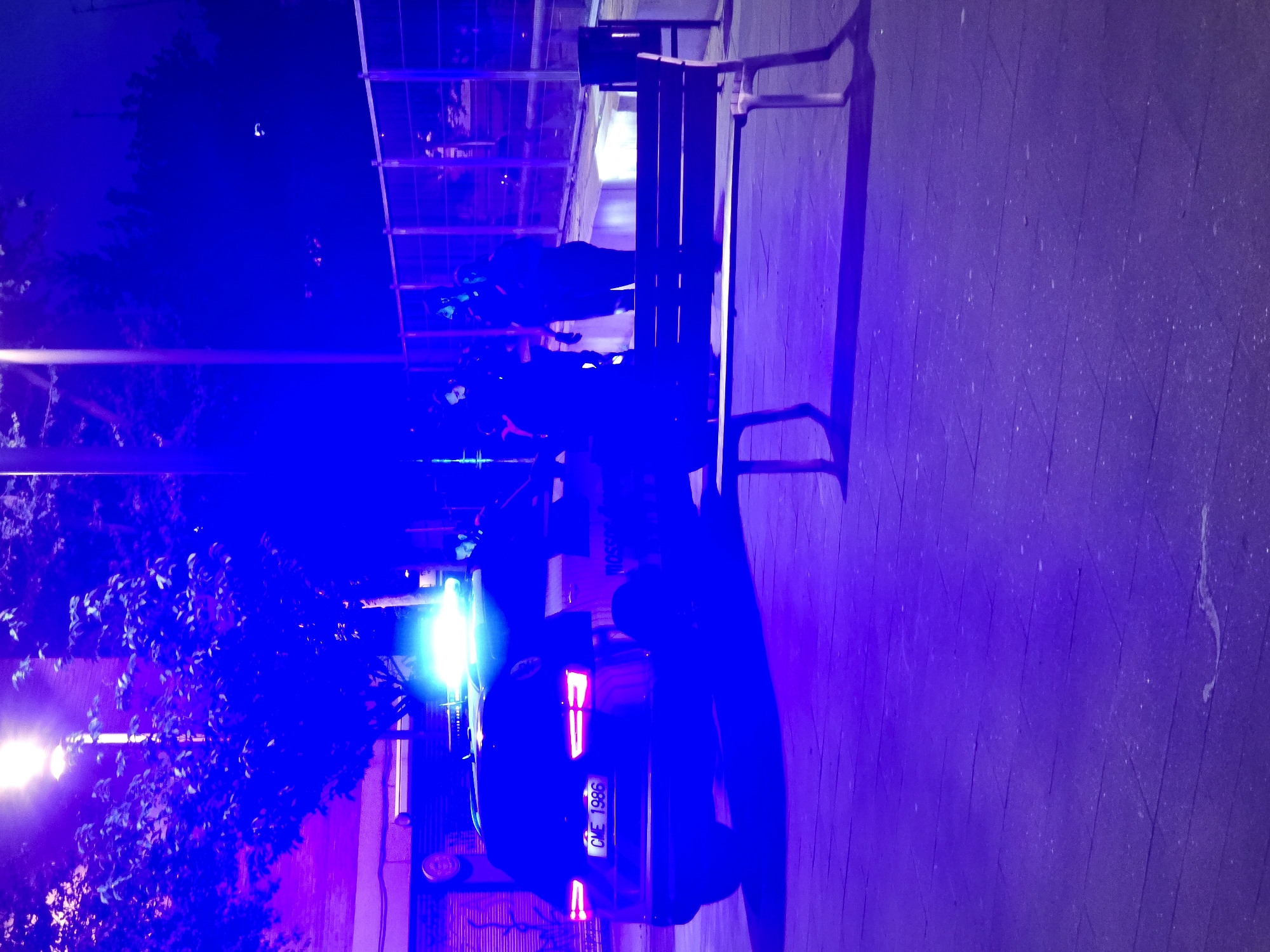 The image, taken at night, displays an outdoor scene illuminated predominantly by strong blue light, indicating an event involving law enforcement. A dark-colored vehicle, likely a police car, is a central focus, positioned towards the bottom right of the frame (when oriented upright). Its rear is visible, featuring illuminated red taillights and intense blue emergency lights that cast a vivid blue-purple hue across the environment. The license plate "CME 1986" is clearly legible on the vehicle's rear.
Several figures, appearing to be people, are visible in the blue-lit scene. Some are positioned near the vehicle, while others are further back, standing or sitting on or near a long wooden bench that extends along the right side of a paved area. Their silhouettes are softened by the low light, making individual details indistinct.
To the left of the vehicle, a large, leafy tree or bush is heavily backlit by both the blue emergency lights and other bright white light sources, possibly streetlights. In the background, a multi-story structure or a tall fence with a grid-like pattern is discernible, also washed in the blue light. The ground is a paved surface, reflecting the intense blue illumination. The overall impression is an active police presence or an incident response in an urban or suburban setting at night, specifically in Cornellà De Llobregat, Spain.