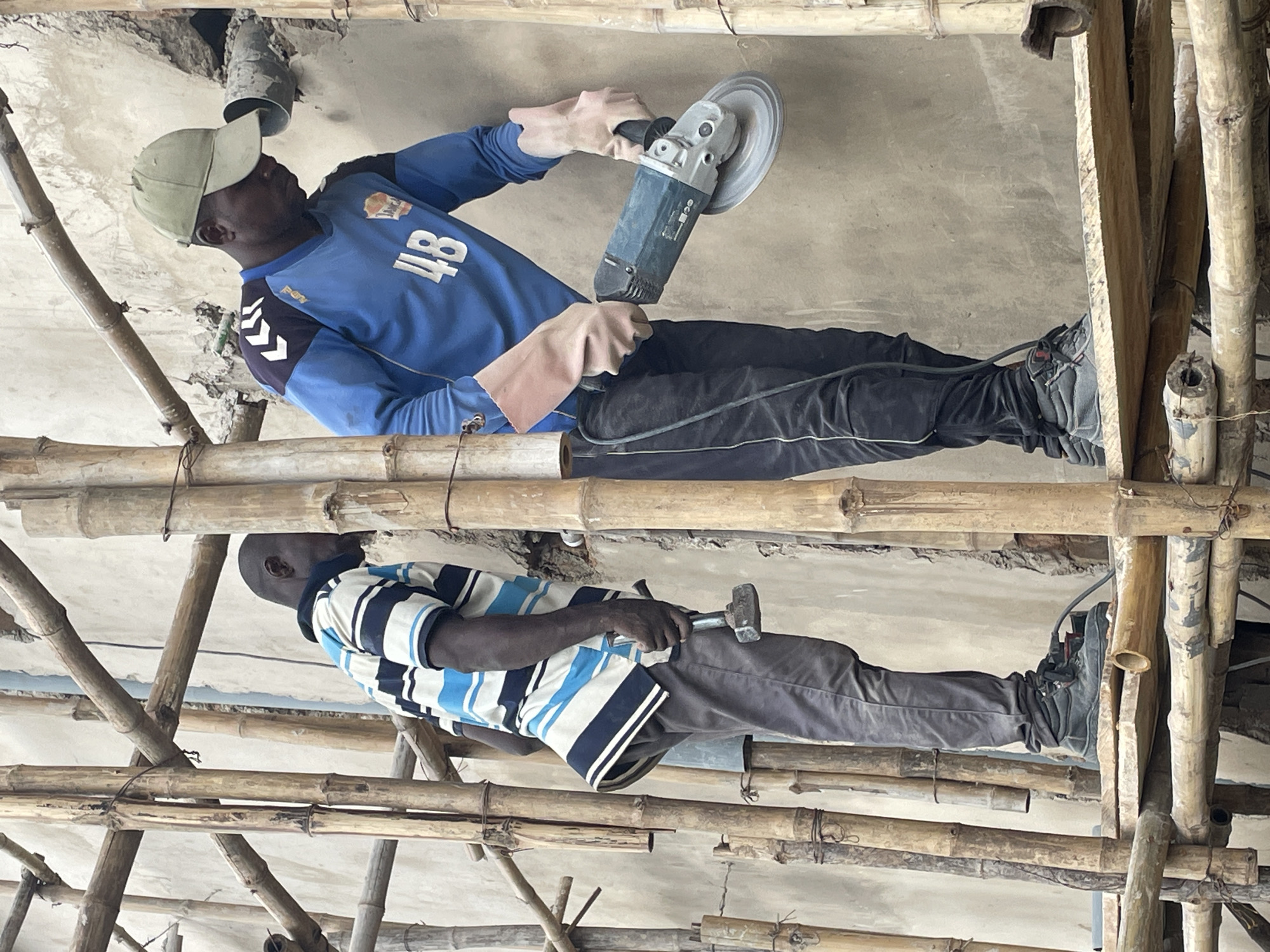 Two men are working on a construction site in Okinano, Nigeria. The man on the left is wearing a striped t-shirt and gray pants, and is holding a hammer. The man on the right is wearing a blue long-sleeved shirt with the number "48" on it and black pants. He is wearing a green cap and protective gloves, and is operating an angle grinder. Both men are standing on a wooden platform supported by bamboo scaffolding, with a concrete wall in the background. The scene depicts an active construction environment.
