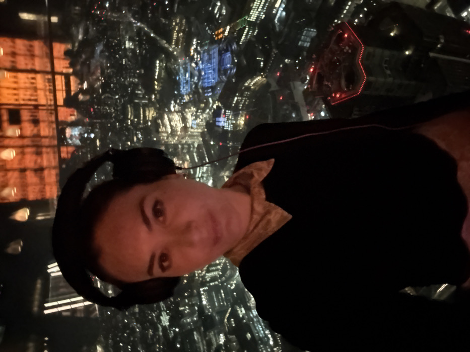 The image displays a female individual in a mid-shot, posing indoors against a nighttime cityscape. The woman faces the camera directly, wearing dark over-ear headphones with a visible cable extending downwards. She is dressed in a dark long-sleeved top with a prominent light-colored, patterned collar. Her dark hair is pulled back, and she has visible earrings.
The background reveals a panoramic view of a brightly illuminated city at night, seen through a large window. Reflections of interior lighting, including hanging fixtures and an orange-toned architectural element, are visible on the glass. The cityscape consists of numerous lit buildings and streets.
The distinct building visible in the lower right of the cityscape, characterized by its zig-zag roofline illuminated with red lights and its classical lower stone architecture, is identifiable as St Andrew Undershaft Church. This vantage point strongly indicates the photograph was taken from a high floor within **The Gherkin (30 St Mary Axe)**, in the **City of London, United Kingdom**.