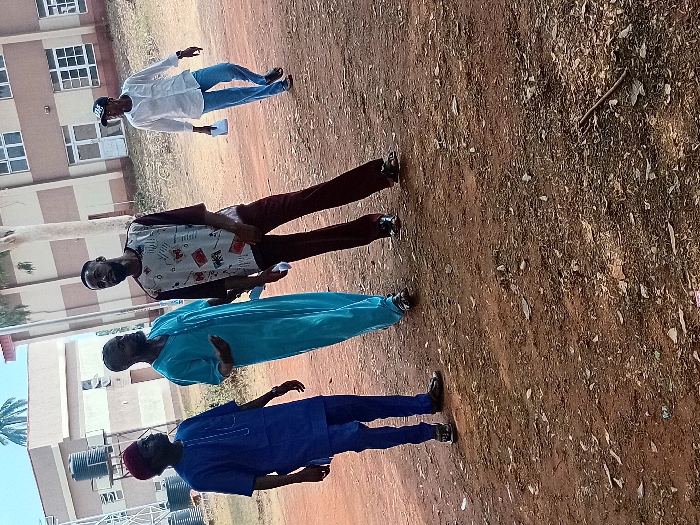 Four men stand on a dirt ground. The man in front wears a blue traditional outfit and a maroon cap. Behind him, another man is dressed in a turquoise robe. The third man dons a graphic t-shirt and brown pants. The last man is dressed in a white shirt and blue jeans, also wearing a cap. The background shows a building with multiple windows and some trees.
