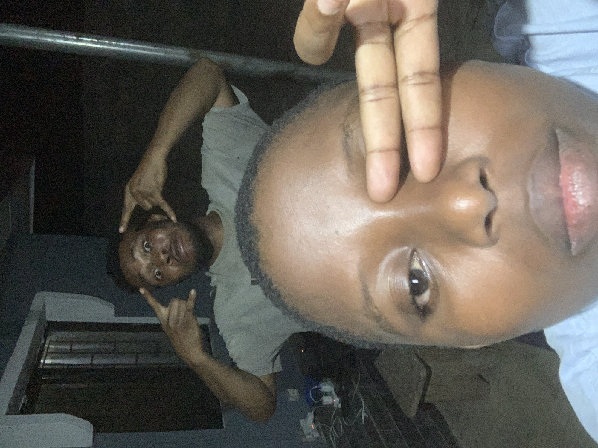 Two dark-skinned men are depicted in an indoor scene. One man is positioned in the foreground with his face close to the camera and his fingers obscuring one eye. He is wearing a light blue collared shirt. Another man stands in the background wearing a grey t-shirt and making a hand gesture. A metal bar runs horizontally across the top of the image. There is a window behind the man in the grey shirt, and electrical cords are visible on the floor. The location is specified as Ikorodu, Nigeria.