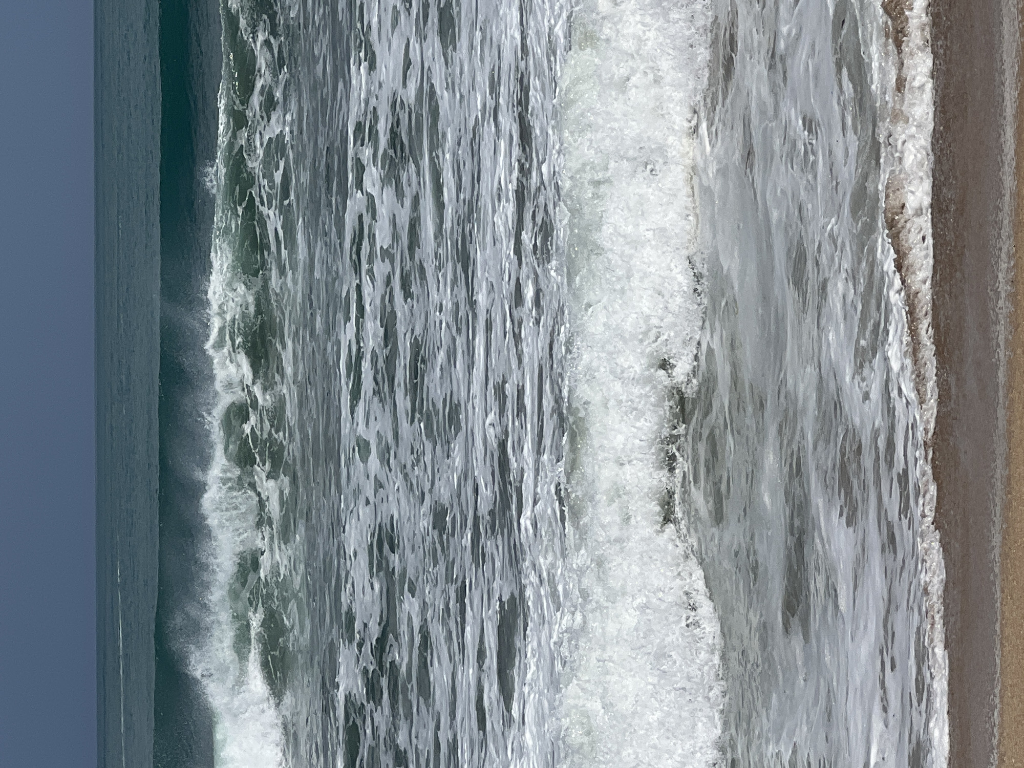 A wide shot shows a choppy ocean with several waves breaking. The water is a deep teal color, with white foam trailing behind the crests of the waves. In the foreground, a sandy beach is visible, wet from the receding tide. The sky above is a dark, overcast grey. This image was taken in Kuta, Indonesia.