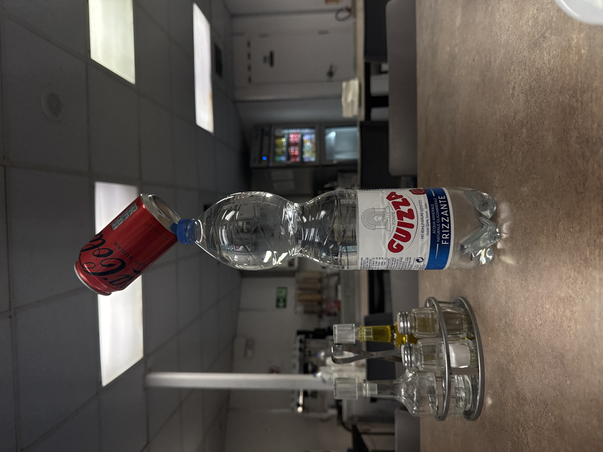 An indoor, eye-level shot captures a still life arrangement on a table, possibly in a breakroom or cafeteria. A red Coca-Cola Zero Sugar can is precariously balanced atop a clear plastic bottle of Guizza sparkling mineral water. The can is tilted, its open end facing downwards, creating a sense of suspended animation. The water bottle, labeled in Italian and featuring a graphic of a classical building, has a blue cap. To the left of the bottle is a metal caddy holding three glass containers: one for salt, one for pepper, and a larger one that could contain oil or vinegar. The table surface is a speckled brown. In the background, a vending machine with illuminated shelves of drinks is visible, along with office-style ceiling lights and a doorway. There are no people present in the image.