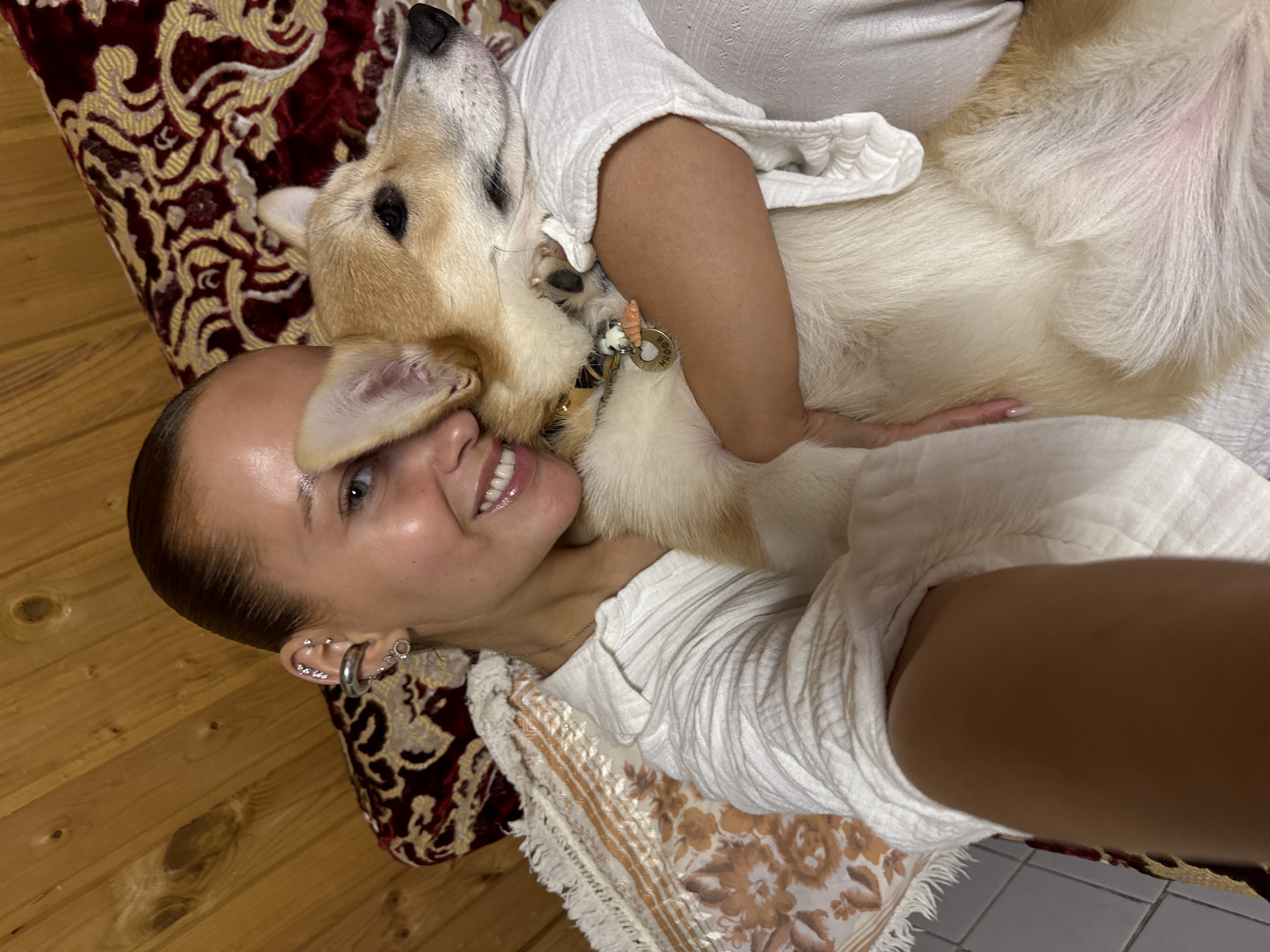 A woman and a dog are lying on a patterned cushion or bed. The woman has fair skin, brown hair slicked back, multiple piercings in her left ear, and is wearing a white, crinkled blouse. She is smiling and looking toward the camera. The dog, which appears to be a Corgi or similar breed, is light brown and white, with its head resting on the woman's face. It is wearing a collar with tags. The background consists of a patterned fabric and a wooden floor. The location in the city of Chastyye, Russia cannot be determined from the image.