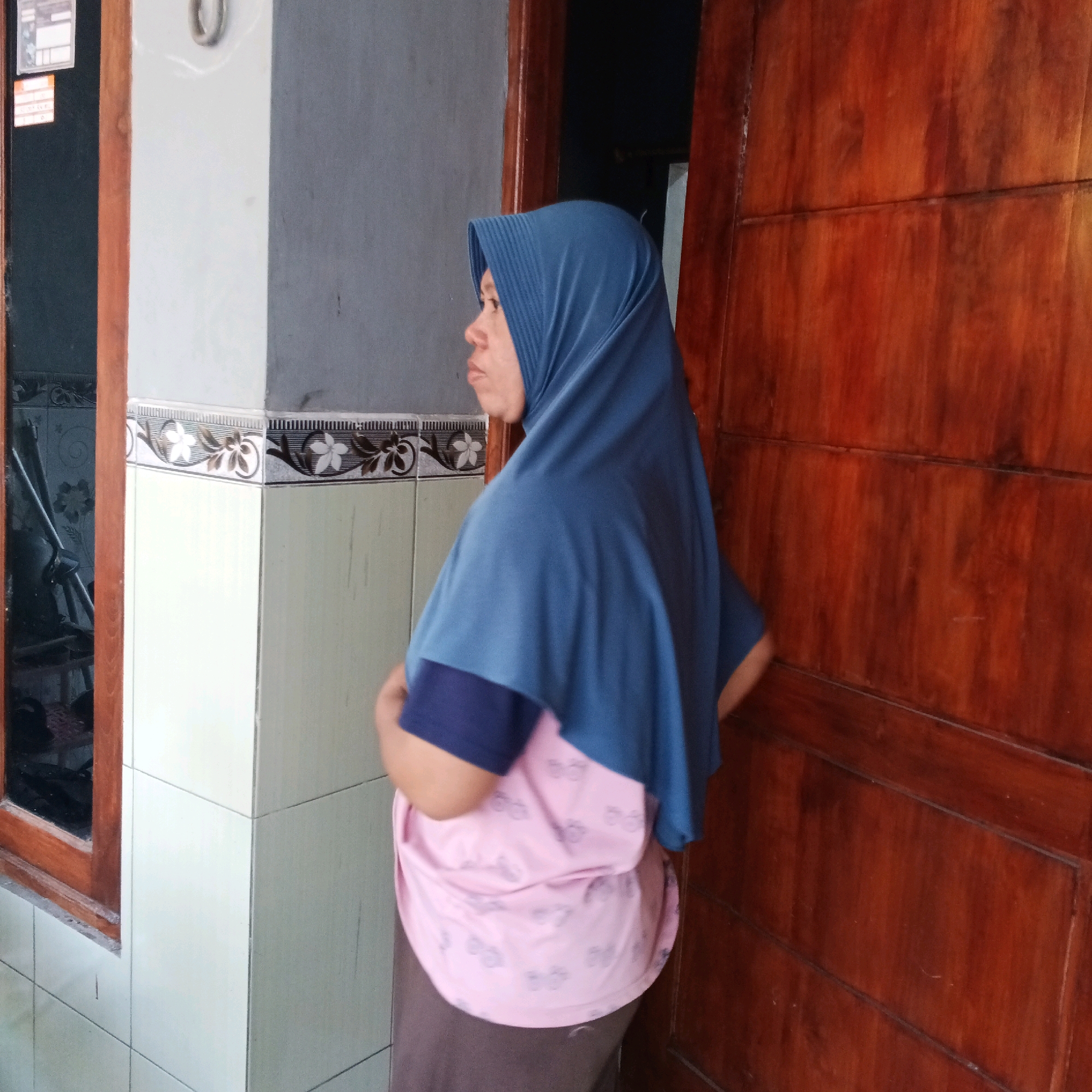 An individual wearing a blue hijab and a pink t-shirt with a pattern of small light purple circles is standing in front of a wooden door. The person's arms are crossed. To the left of the individual is a wall tiled with square white tiles. Above the white tiles is a decorative border with a floral pattern. Further to the left is a window with a wooden frame. The overall setting appears to be an outdoor or semi-outdoor space.
