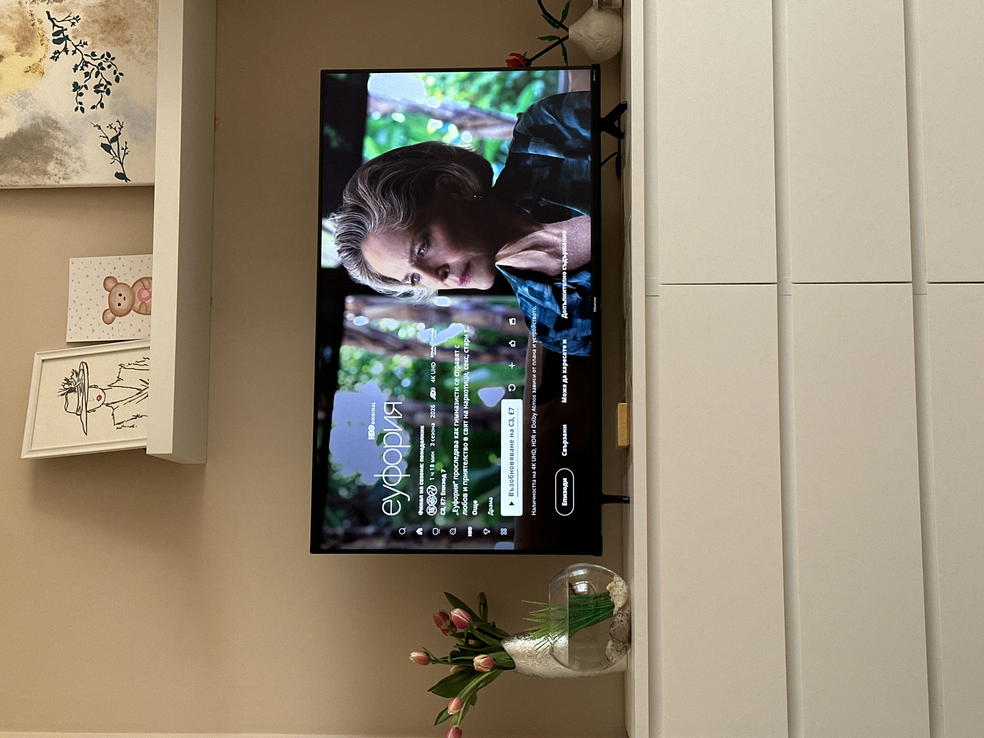 A vertically-oriented interior shot shows a television mounted on a wall between a shelving unit and a set of cabinets. The television screen displays an episode of the HBO series "Euphoria." The episode is listed as "S3, E7: Episode 7." The characters on the screen are not clearly visible. To the left of the television is a partially visible shelving unit with a framed abstract painting at the top, followed by a framed cartoon bear, and a framed line drawing of a person in a hat. Below the television is a vase with pink tulips and green foliage. To the right of the television is a large white cabinet with grooved panels. The wall is painted a light beige color. Based on the Cyrillic text on the television screen, the location is likely Bulgaria. Without more specific information, I cannot pinpoint the location in Sofia.
