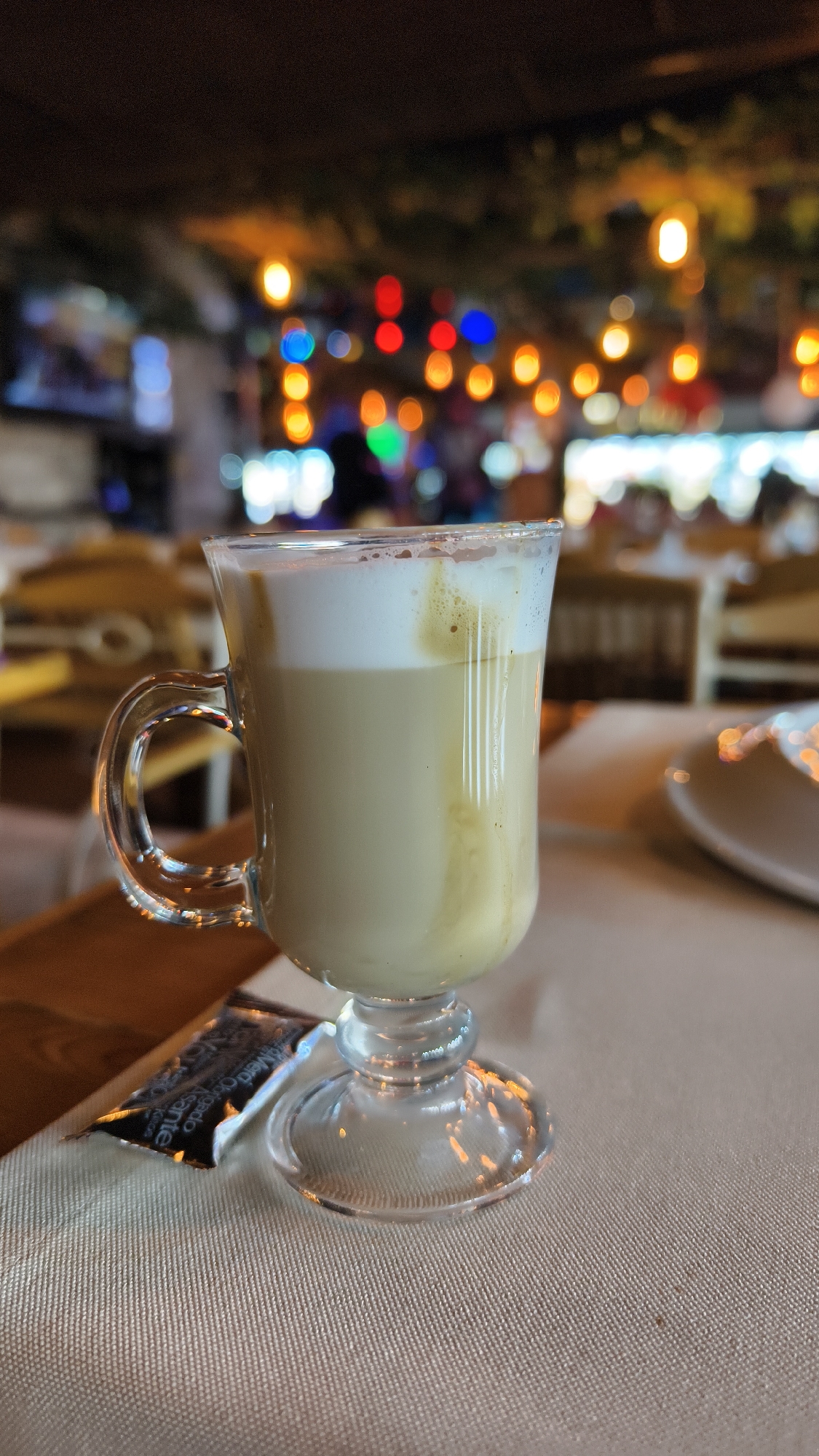 An eye-level, medium shot captures a glass of latte on a textured beige tablecloth in what appears to be a dimly lit restaurant or cafe. The glass, with a handle, contains a light brown latte topped with a frothy white layer. To the left of the glass, a dark packet, possibly containing sugar or creamer, is partially visible. The background is out of focus, displaying a bokeh effect of colorful lights in reds, blues, yellows, and white, suggesting a vibrant evening atmosphere. In the mid-background, blurred shapes of chairs and tables are discernible, hinting at a dining setting. The overall impression is one of a cozy, possibly upscale establishment, typical of popular tourist spots like Kuşadası, Turkey.