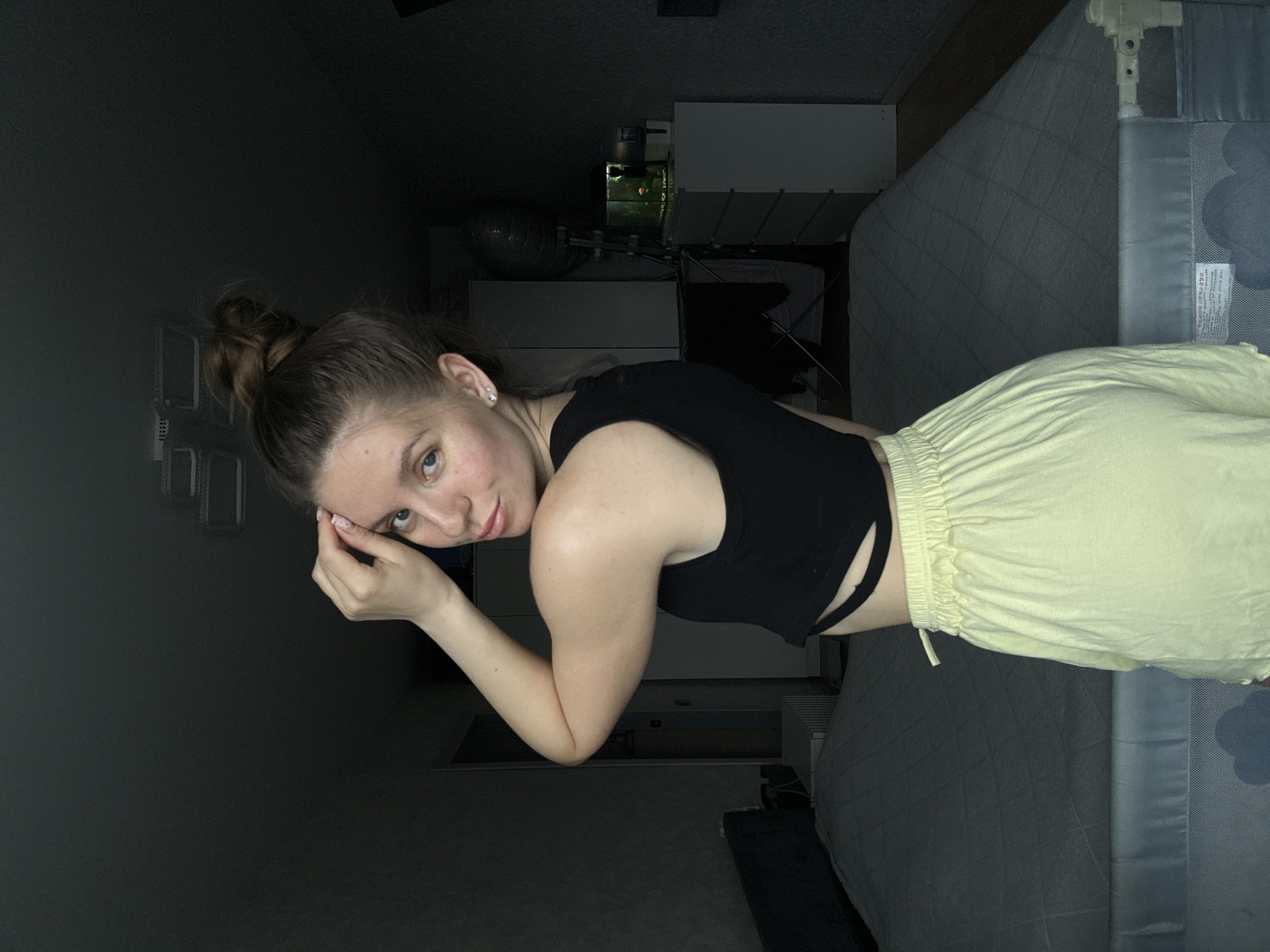 A young woman is depicted in a dimly lit indoor setting, likely a bedroom. She is seen from the waist up, lying down or reclining on a bed, looking directly at the camera with a calm, soft smile. She has fair skin, light-colored eyes, and her brown hair is styled in a bun on top of her head. She wears a black sleeveless crop top and light yellow, loose-fitting pants or shorts with a drawstring waist. A small stud earring is visible in her left ear, and her left hand is raised near her temple.
The background is dark and somewhat indistinct due to the low lighting. To the right of the woman, a grey quilted mattress and a white crib railing with a light blue cloud pattern are visible, indicating the presence of a child's sleep area. A small, white tag with illegible text is attached to the crib railing. Further in the background, various pieces of dark furniture, possibly chests of drawers or shelves, can be discerned. A small, illuminated aquarium or similar object is vaguely visible in the distant background. The overall subdued lighting suggests it is either evening, night, or a room with closed curtains. The image does not contain specific visual cues to confirm the stated location of Klin, Russia.