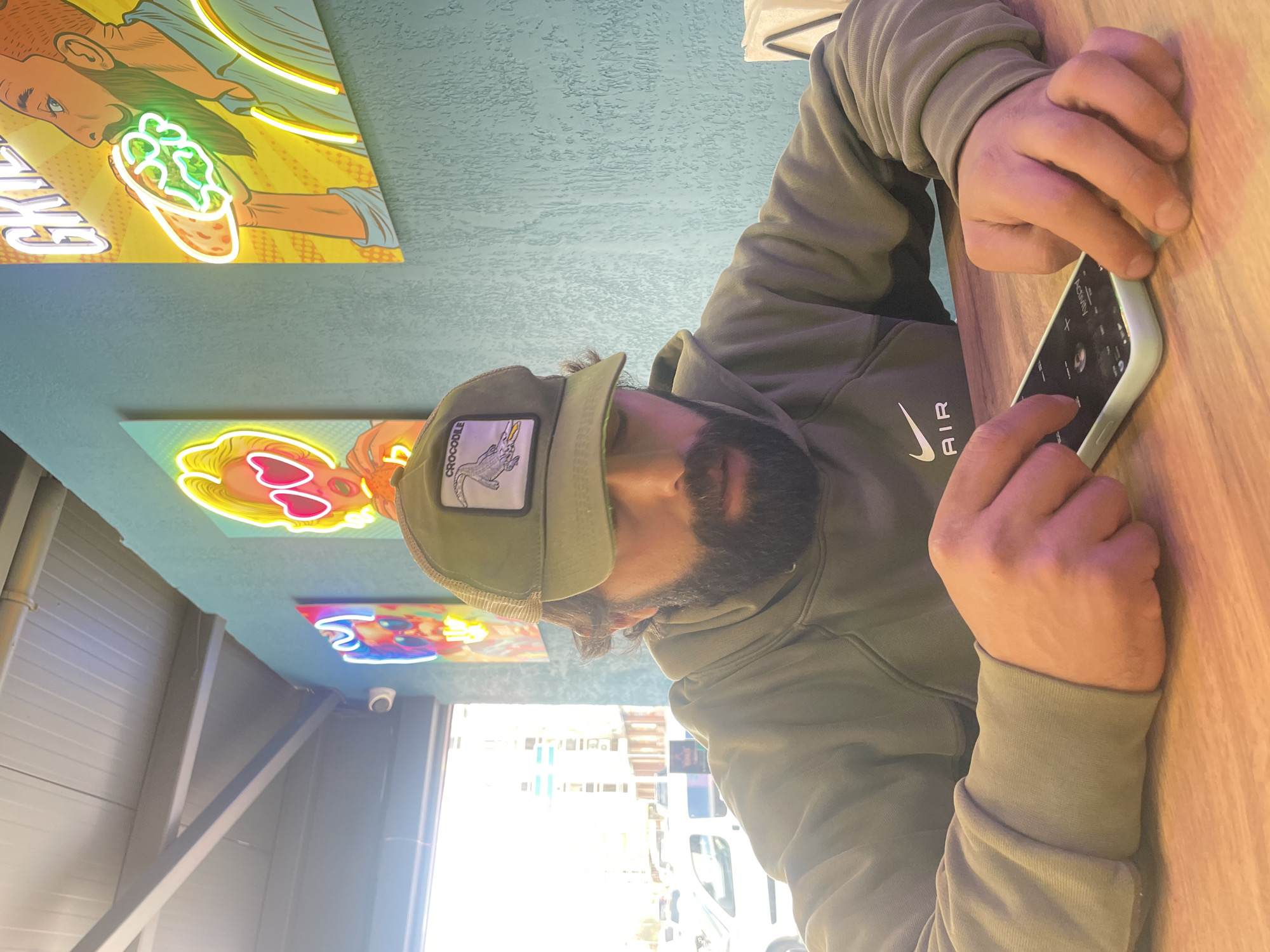 A man with a beard wearing an olive green hoodie with the word "AIR" and a white Nike logo on the right side of his chest, along with an olive green baseball cap with a crocodile patch, is sitting at a light brown wooden table. He is holding a smartphone in a light green case with his right hand. Above him on a blue textured wall are various neon signs, including one of a cartoon man holding a food item, and another of a cartoon woman with heart-shaped sunglasses. Outside the window, visible through a partially obstructed view due to the bright daylight, there appear to be buildings and parked vehicles, indicating an urban setting. The location is identified as Krasnoyarsk, Russia.
