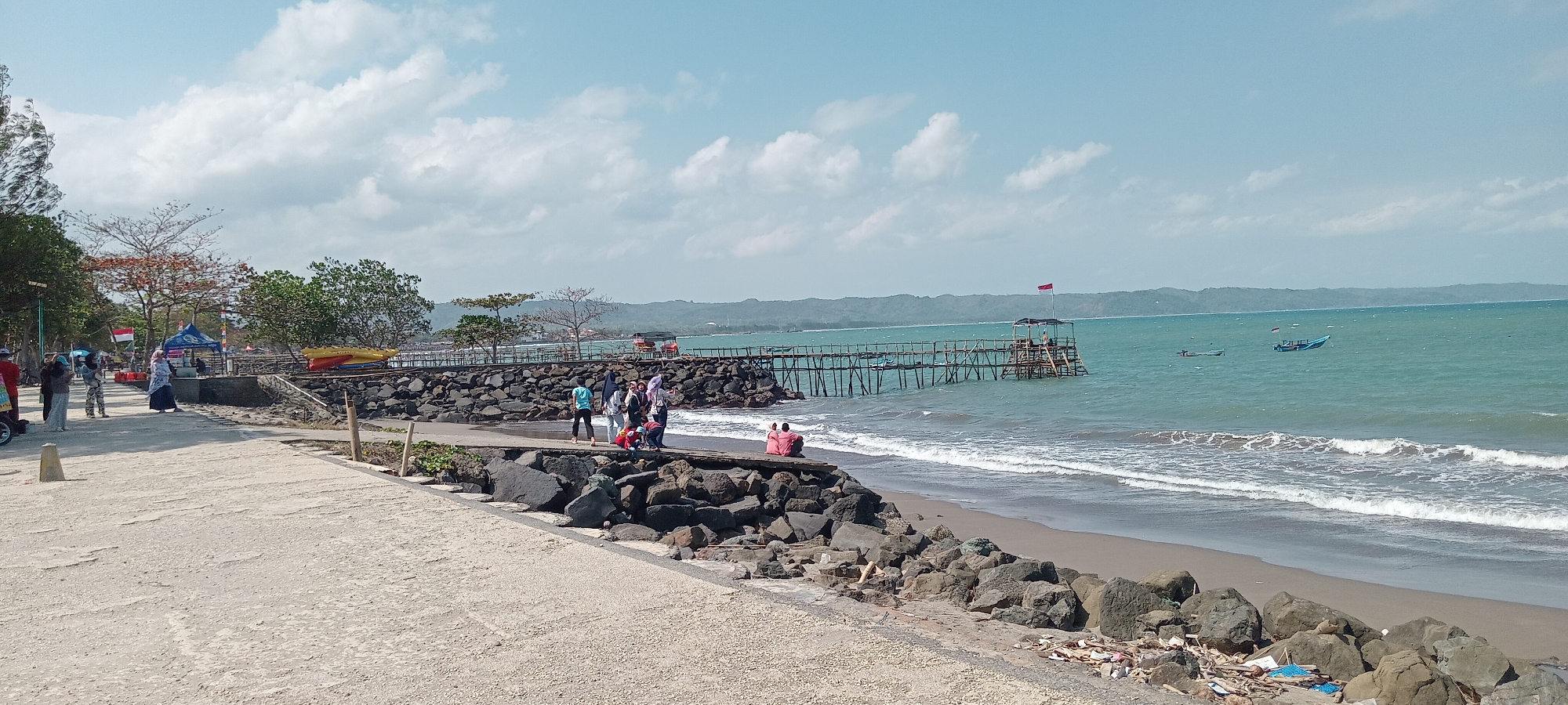 The image depicts a seaside scene in Desa Pagandaran, Indonesia, under a partly cloudy sky. In the foreground, a paved pathway runs along the coast, bordered by a rocky embankment. Several individuals are seen on this pathway. To the right, the sea meets a sandy beach, with waves rolling ashore and a line of rocks along the water's edge. Further out, a wooden pier extends into the turquoise water, featuring a small structure on its end topped with a red flag. Two small boats are visible in the distance. Along the left side of the image, trees and some temporary structures, including tents, are present, suggesting a recreational area. A group of people is gathered on the pathway near these structures.