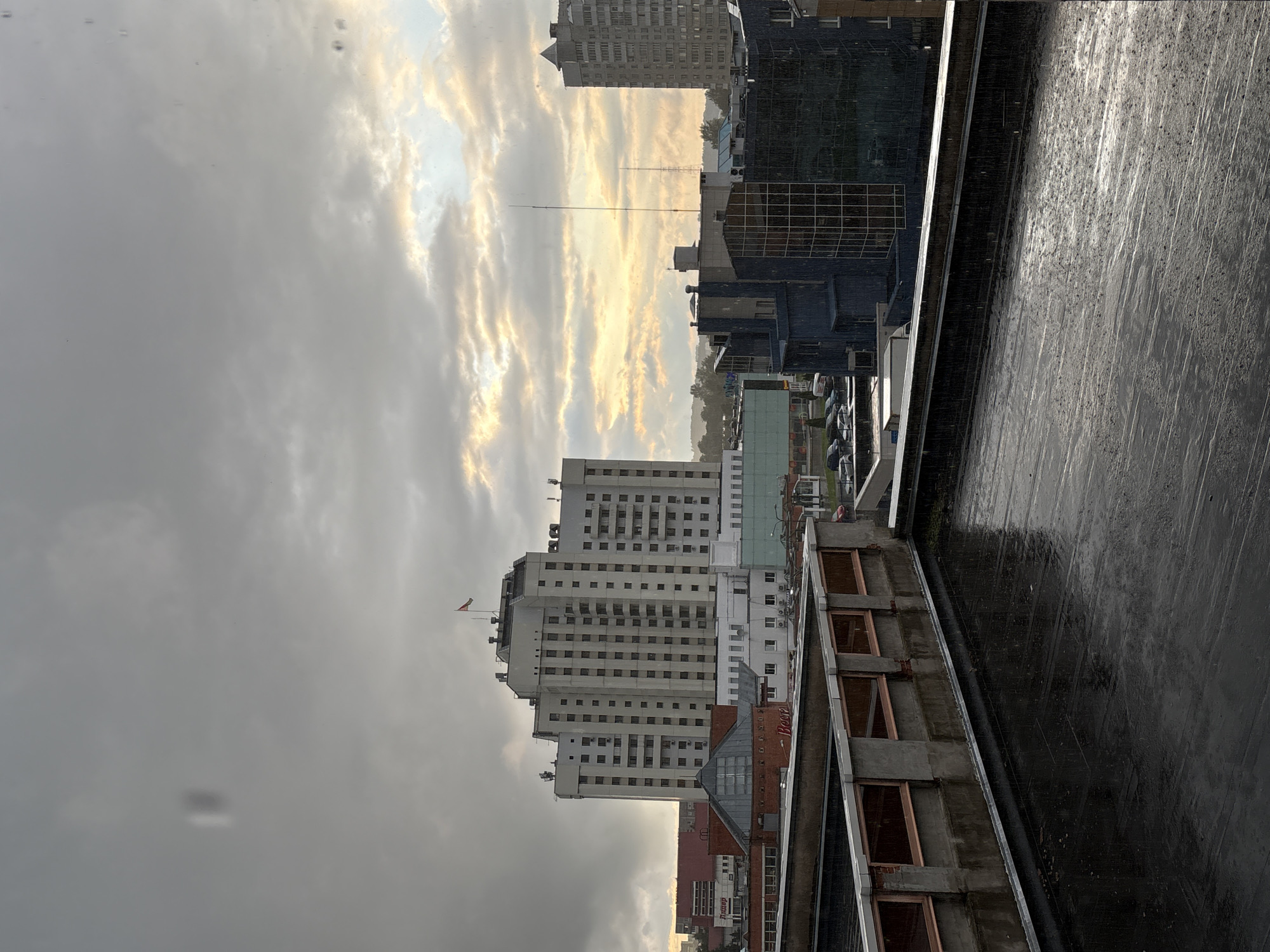 The image depicts an overcast sky with patches of sunlight breaking through clouds, suggesting a cloudy or rainy day. In the foreground, a wet, dark-colored rooftop, likely made of asphalt or a similar material, shows reflections and small puddles, indicating recent or ongoing precipitation. A concrete structure with openings lined with copper-colored material runs along the edge of the rooftop.
In the mid-ground, a large, multi-story building with a light-colored facade and numerous windows dominates the scene. A flag, possibly the flag of Belarus, is visible on the rooftop of this building. Several other buildings of varying heights and architectural styles are also present, including a darker, modern building with a glass facade, and a residential-style high-rise. Various vehicles are parked in a lot between the buildings.
The scene appears to be an urban environment, specifically the city of Vitsebsk, Belarus, given the architectural style and the presence of the Belarusian flag. The overall impression is one of a somewhat dreary, wet day in a city. There are no discernible people or active interactions captured in the frame. The weather conditions, characterized by rain and clouds, are a prominent feature of the scene.