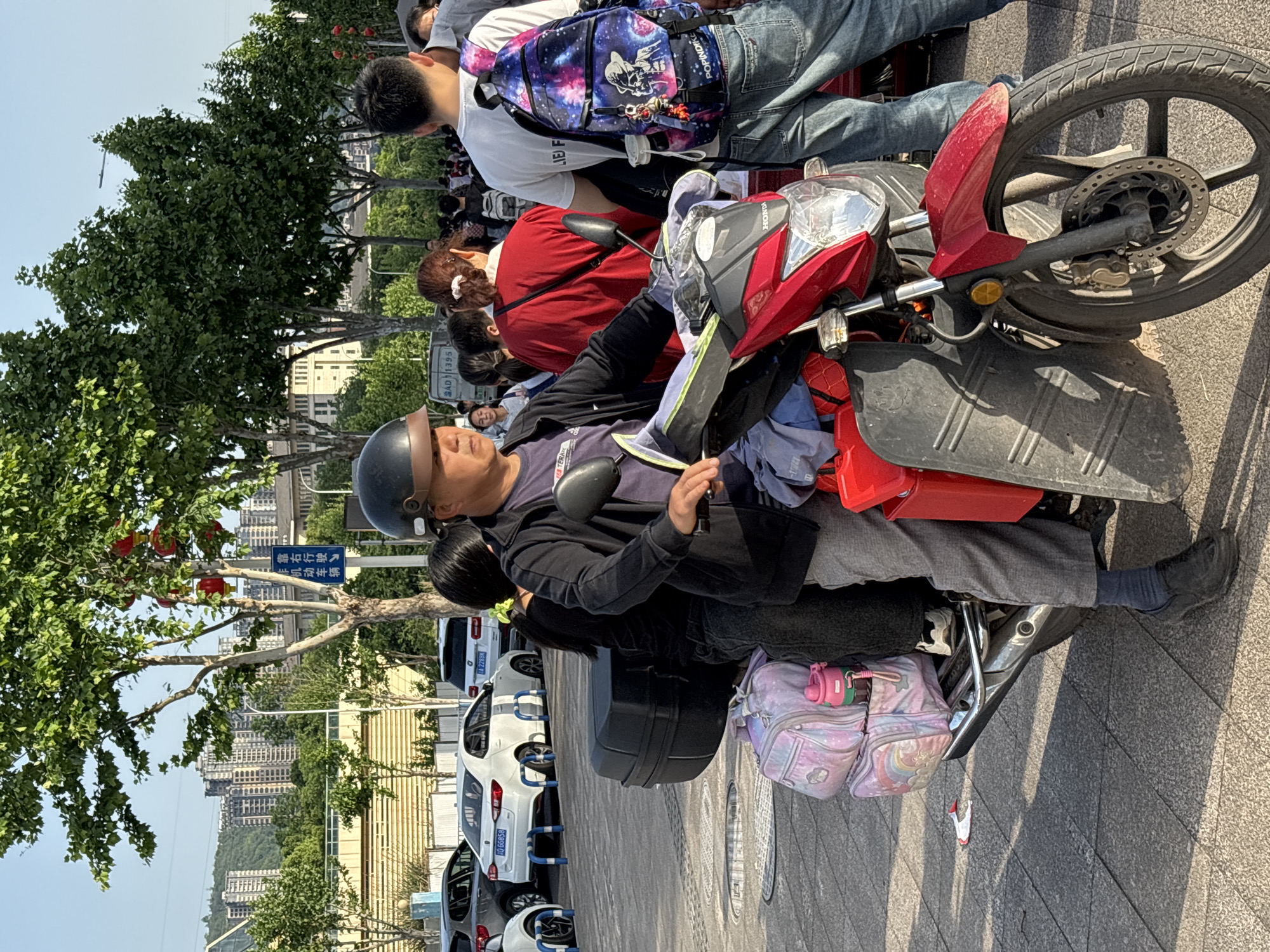 A man wearing a helmet is seated on a red scooter. He is looking down at something in his hands. Behind him on the scooter sits a small girl with black hair. The scooter has a large black storage box and a pink backpack. Behind the scooter is a line of parked white cars. On the left side of the frame, there are trees and red lanterns hanging. A blue street sign is partially visible with Chinese characters. In the background, there are several tall buildings, which suggest this is a city in China. Based on the license plate numbers, the location is Jiangjin, China. Many other people are in the background.