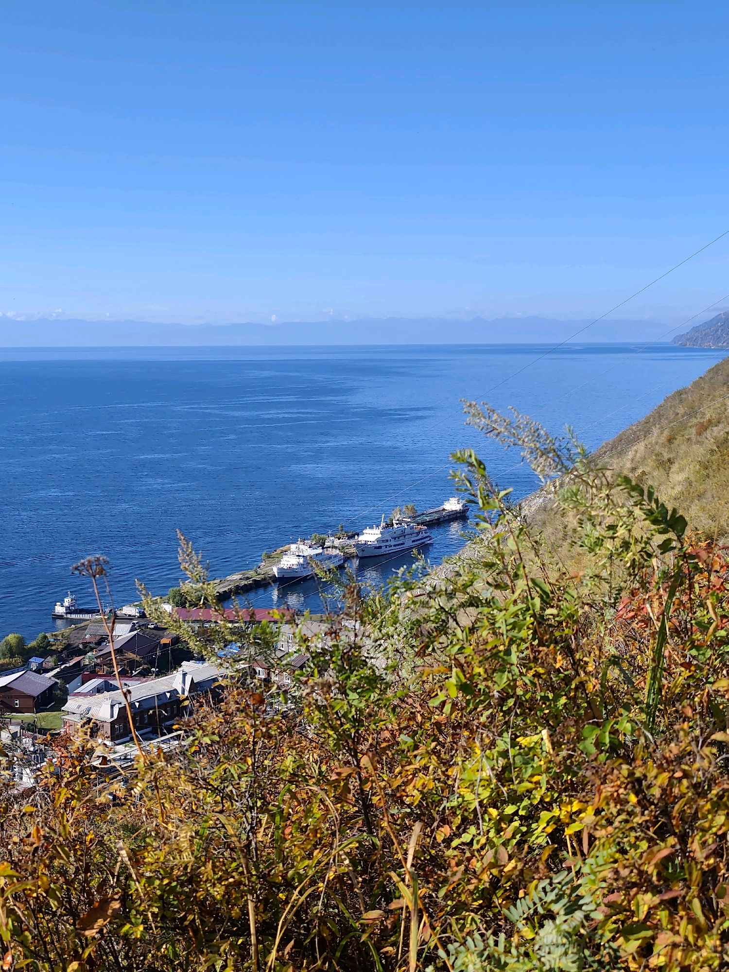 This image captures a scenic view of a port town on the shores of Lake Baikal, Russia, on a clear, sunny day. In the foreground, lush autumnal foliage in shades of green, yellow, and brown partially obscures the view, creating a natural frame. Beyond the foliage, a cluster of buildings with varied roof colors, predominantly red and brown, forms a small settlement. A pier extends into the deep blue waters of the lake, where several white passenger ferries are docked. The lake stretches out to the horizon, where a faint line of mountains under a vast, clear blue sky can be seen. The overall impression is one of serene natural beauty and the tranquil activity of a lakeside community.