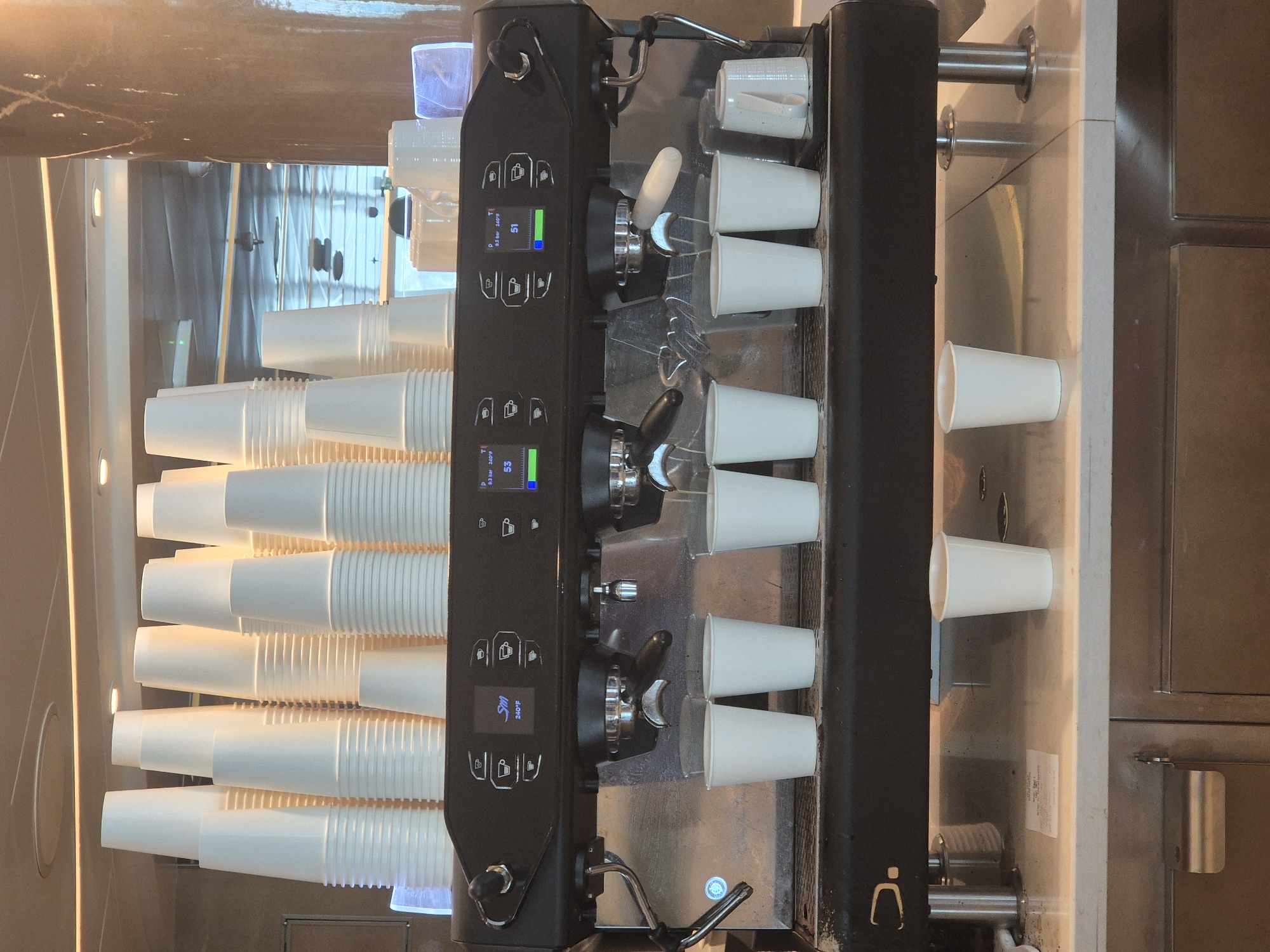 A close-up, top-down shot reveals a black, commercial espresso machine laden with white paper cups, set against a backdrop of metallic and neutral-toned surfaces. The espresso machine is flanked by stacks of cups on the left, and individual cups are held in place by the machine on the right. Control panels on the espresso machine display digital readouts, and steam wands are visible. The environment appears to be a modern kitchen or commercial food service area. There is no direct evidence linking the location to Rapallo, Italy.