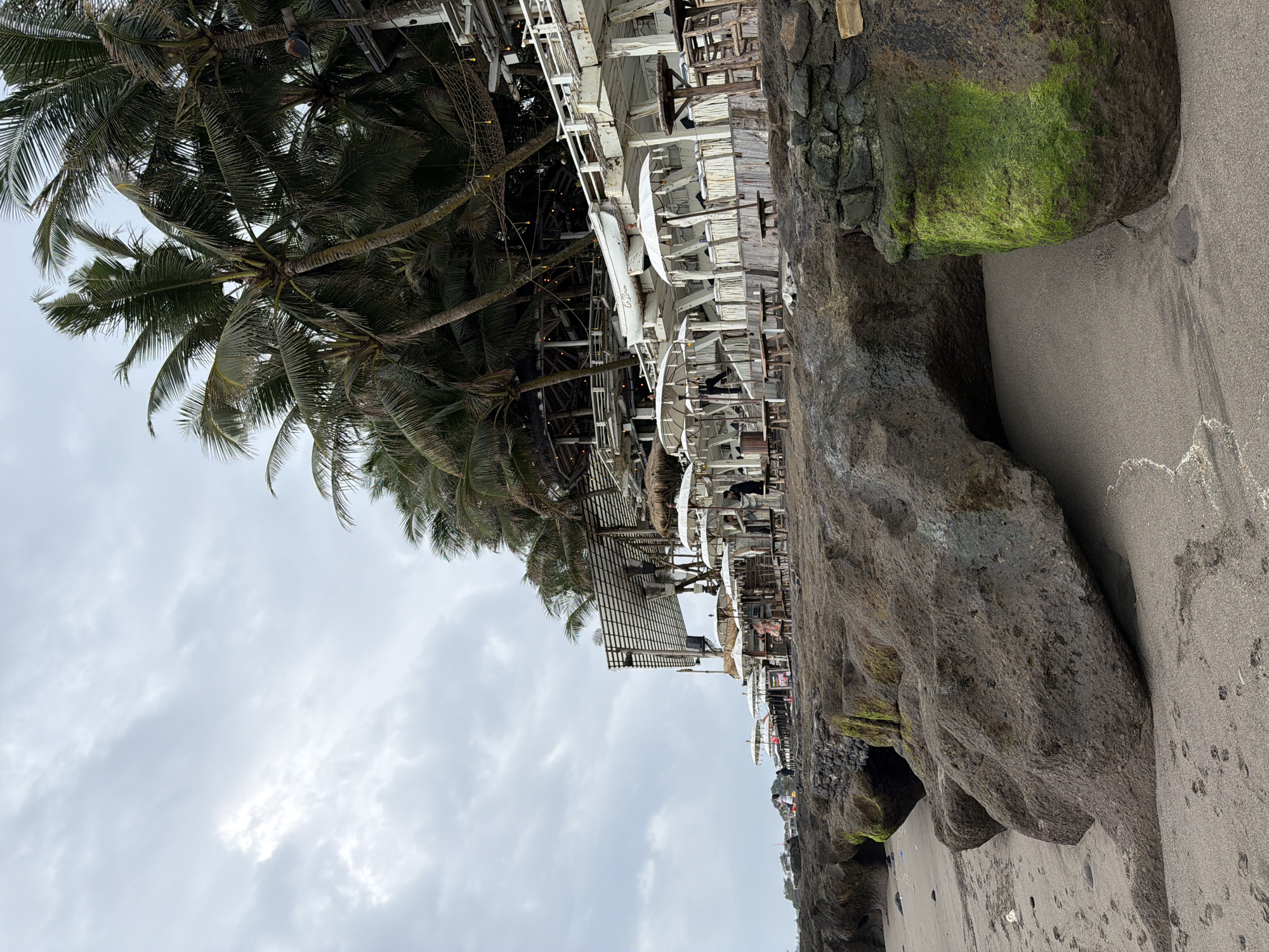 A view of the beach and restaurant in North Kuta, Indonesia, on a cloudy day. The sand is dark grey, with footprints visible. Large, dark grey boulders, some covered in bright green moss, are scattered on the beach. Behind the rocks, a beachside restaurant with white umbrellas and wooden seating is visible. Palm trees with dark green fronds line the top of the restaurant structure. Several people are seen at the restaurant and on the beach. The sky is overcast with light grey clouds.