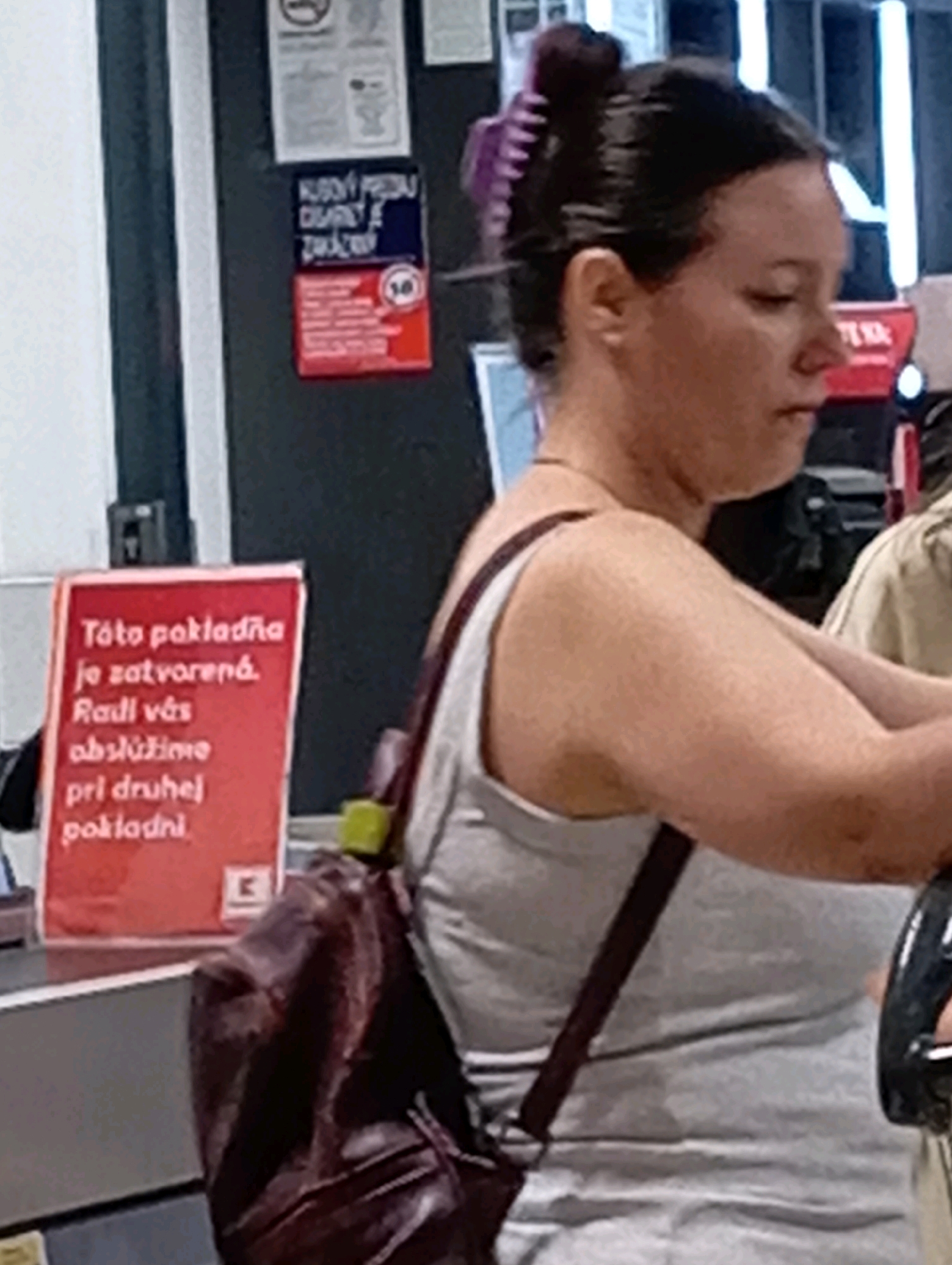 A woman is standing in a store in Nitra, Slovakia. She has her dark hair pulled up in a bun with a purple clip. She is wearing a gray tank top and a dark brown backpack is slung over her shoulder. A red sign is in the foreground, to the left of the woman. The sign has white text that says "Tato pokladňa je zatvorená. Radi vás obslúžime pri druhej pokladni." Below the text, there is a white 'K' logo in a square. Behind the woman, there are several signs on the wall. One sign is red with white text and a circle with a line through it, indicating a prohibition. Another sign above it is blue and white. The overall environment appears to be a checkout area in a retail store, possibly a supermarket.