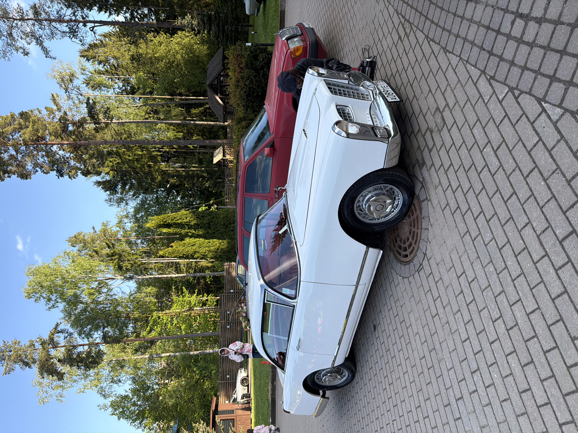 The image depicts two vintage cars parked on a cobblestone street in Repino, Russia. A white classic car with chrome accents is in the foreground, bearing the license plate A089TE 98. Adjacent to it is a red car, with a person wearing dark clothing leaning into its engine compartment. The background features a wooded area with tall trees against a blue sky. A person is standing near a small wooden structure in the distance. The scene appears to be a vintage car event or gathering.