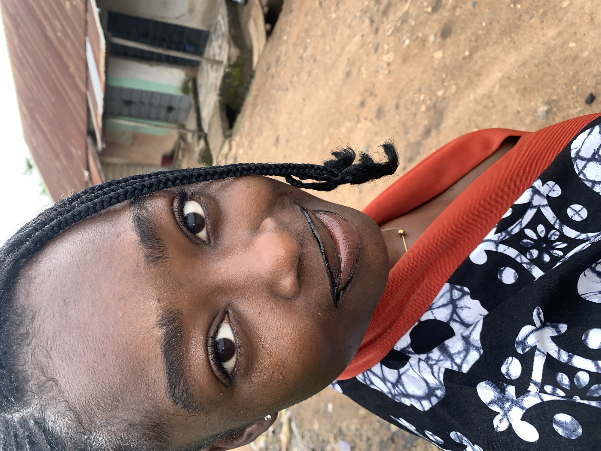 A close-up, high-angle shot captures a young Black woman from the chest up, posing outdoors in what appears to be a rural or informal urban setting in Rafin Par, Nigeria. She has dark skin, dark eyes, and black braided hair, with one braid falling across her face. She is wearing a black and white patterned garment with a bright orange collar and a thin gold necklace. Her lips are painted with black lipstick. In the background, a modest, weathered building with a corrugated metal roof is visible. The ground in front of the building is unpaved and appears to be dirt or gravel. The overall scene suggests a casual, everyday moment in a Nigerian village or settlement.