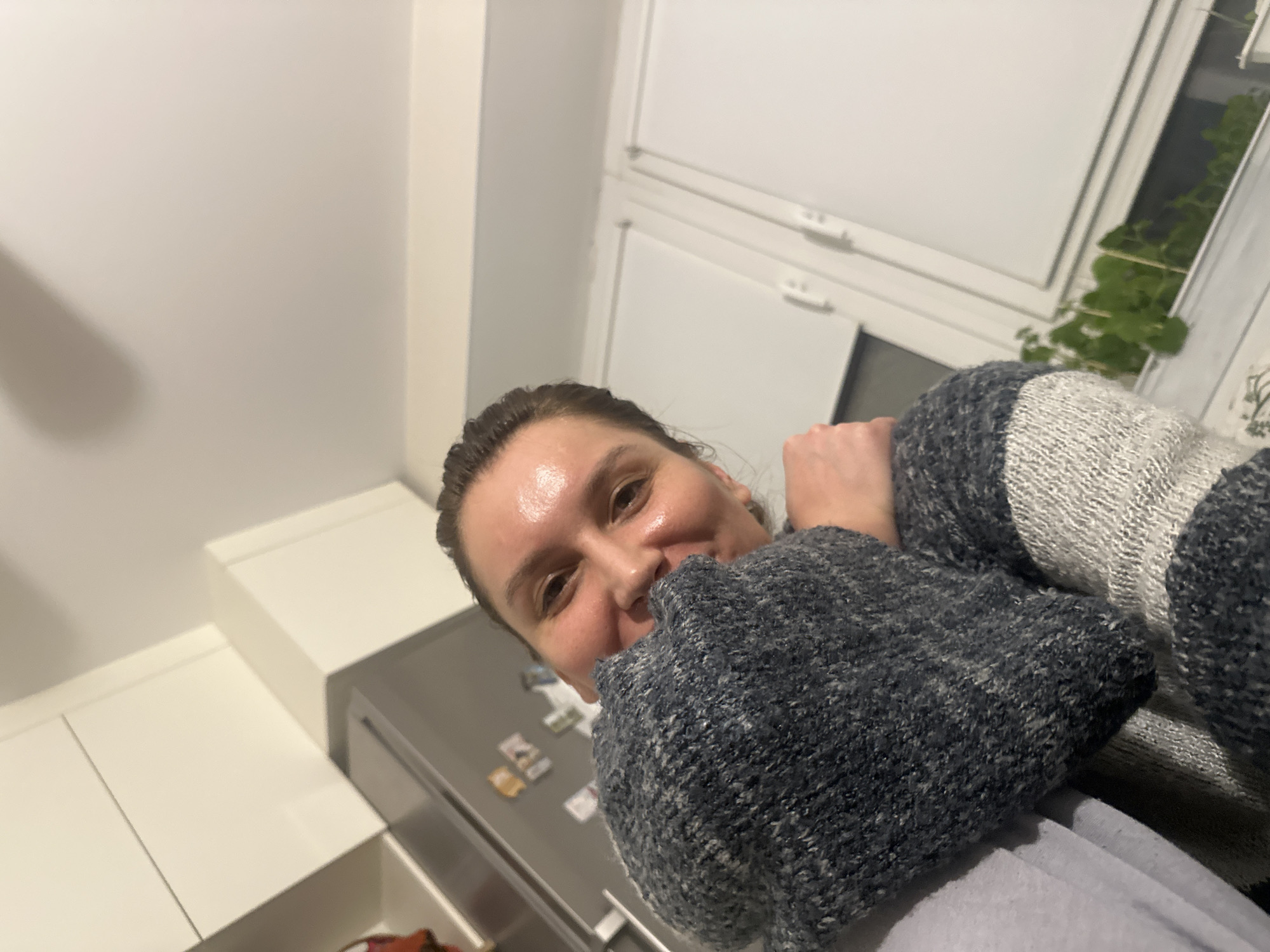 The image captures a cheerful adult taking a low-angle selfie or portrait in what appears to be a modern kitchen setting in Moscow, Russia. The person, with brown hair pulled back, smiles broadly at the camera, their face illuminated with a slight sheen on the forehead and cheeks. They are wearing a thick, knitted sweater featuring a pattern of dark grey and light grey/white, with their arms tucked comfortably within the sleeves.
In the background, a contemporary kitchen layout is visible. To the left and below the person are white, flat-front cabinets or drawers, reflecting a clean aesthetic. A prominent grey or stainless steel refrigerator stands behind the individual, adorned with several small, rectangular magnets or notes that are indistinct due to blur. Above and to the right, white-framed windows or blinds are visible, with a vibrant green potted plant peeking through one of the panes, adding a natural element to the indoor scene. The lighting suggests an interior setting, possibly artificial, indicating it could be evening, night, or an overcast day. The overall impression is one of warmth and contentment in a cozy home environment.