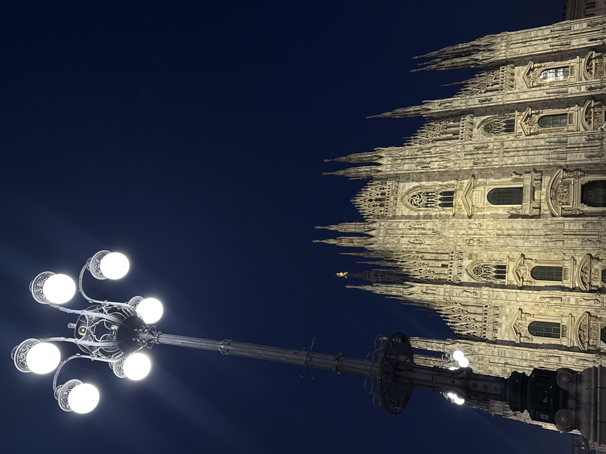 The image depicts the Duomo di Milano at night, viewed from a low angle. The facade of the cathedral is illuminated. A lamppost with multiple spherical lights is prominently featured in the foreground, contrasting with the intricate architectural details of the Duomo. The sky is a deep blue.