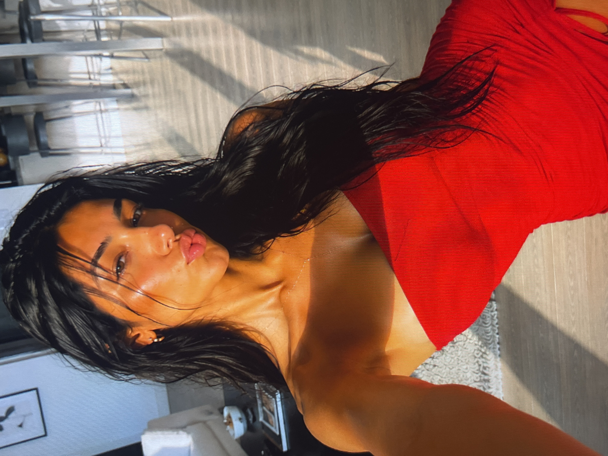 The media file shows a woman in a red dress, facing the camera. She has dark hair and is wearing makeup. Her lips are slightly parted, and she appears to be posing. The lighting suggests it is daytime, with shadows cast across the floor. The background is blurred but appears to be an indoor setting with some furniture. The overall tone of the image is intimate and personal, possibly a selfie. The woman's expression is somewhat playful or alluring.