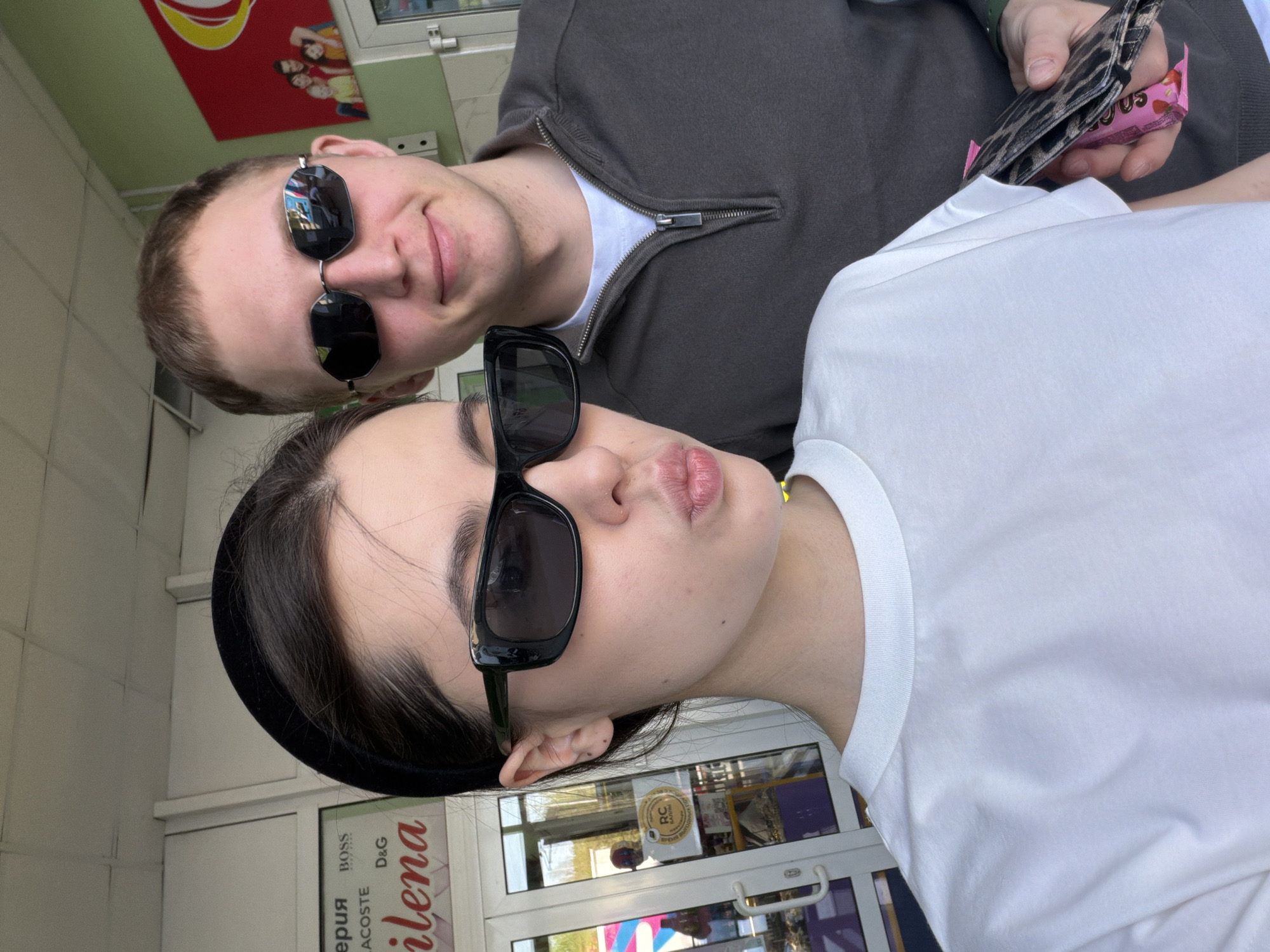 A selfie shows two people, a man and a woman, likely standing in front of a store. The woman is in the foreground wearing a white t-shirt and a black headband. She has dark hair and is wearing black sunglasses, pursing her lips in a pout. The man is behind her, wearing a brown zipped jacket over a white shirt and sunglasses. He is holding a wallet and a pink package. The scene is outdoors, with the entrance of a store visible behind them. The door of the store has lettering, and some of it is identifiable as "Lena" vertically written on the door. The door and sign are for a shop in the city of Chaikovsky, Russia.