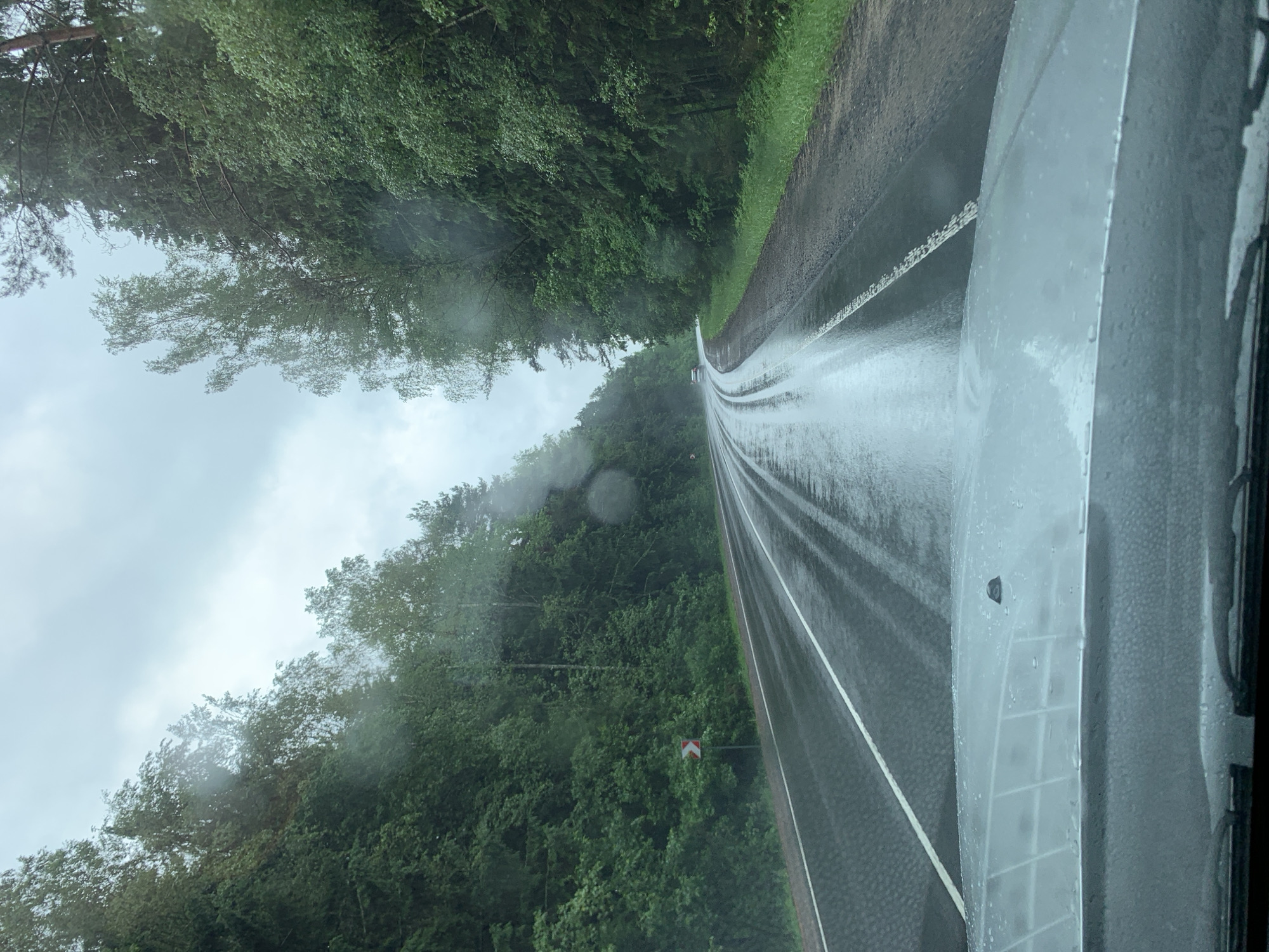 The image depicts a wet, paved road with a white line marking its center, viewed from the perspective of a vehicle's dashboard. The road curves to the left, bordered by dense green trees on both sides, with a cloudy sky overhead. Raindrops are visible on the windshield, and the road surface is glistening with water, indicating recent or ongoing precipitation. A red and white directional sign is visible on the left side of the road. A distant car is seen on the road ahead. The scene appears to be a rural or forested area in the Сваткаўскі сельскі Савет, Belarus, given the natural landscape and the type of road.