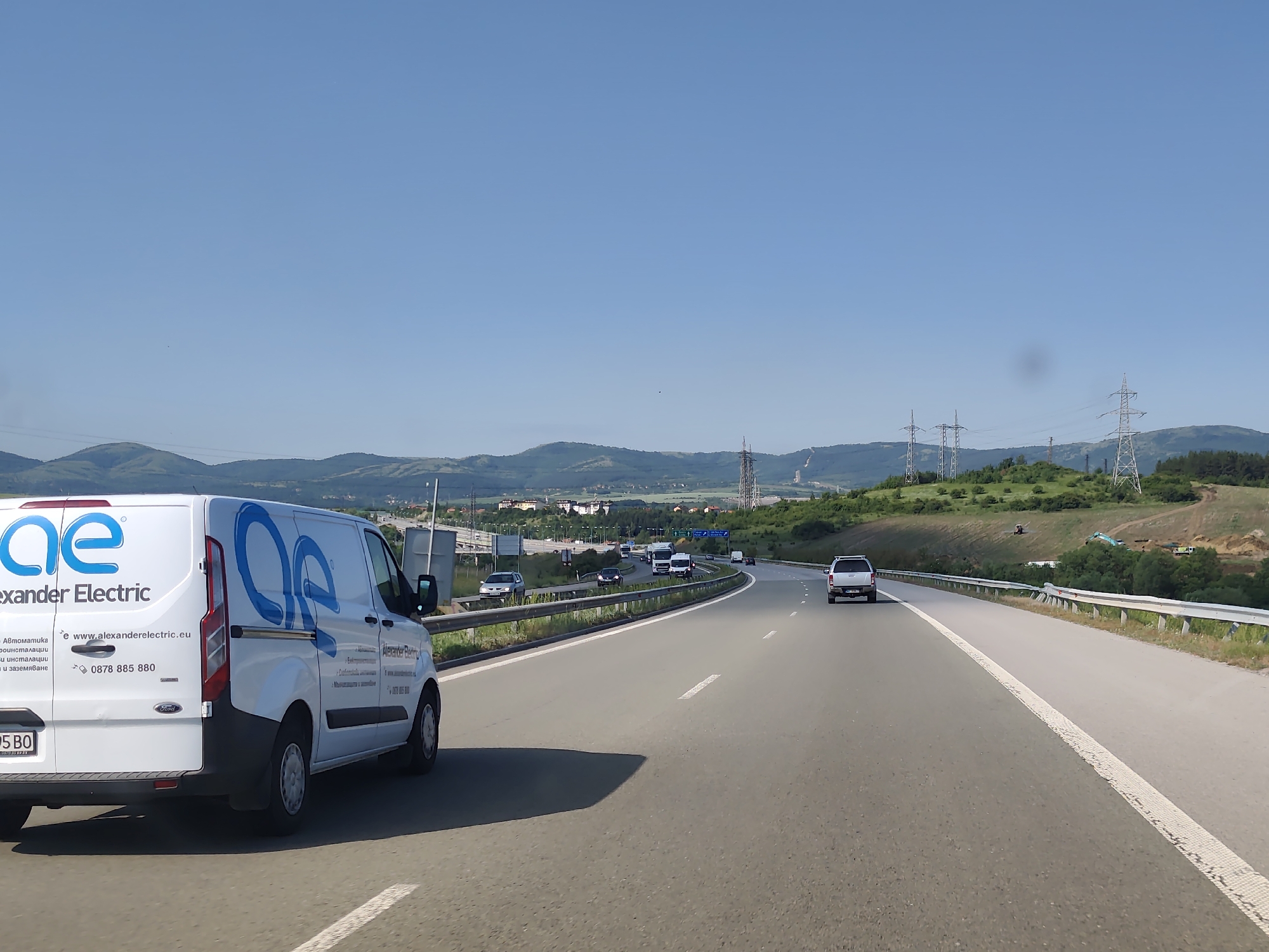 The image captures a highway scene near Dragichevo, Bulgaria, on a clear day with a bright blue sky. In the foreground, a white van with the logo "oe Alexander Electric" is driving on the right side of the road. The van is branded with text that includes a website address and a phone number. Several other vehicles, including cars and trucks, are visible on the multi-lane highway. The road is bordered by a metal guardrail and a grassy embankment. In the background, rolling hills covered in green vegetation and trees are visible, interspersed with power lines and electrical towers. Some construction activity, including earthmoving equipment, is present on the hillsides. The overall scene depicts traffic on a sunny day along a highway surrounded by natural and developed landscapes.