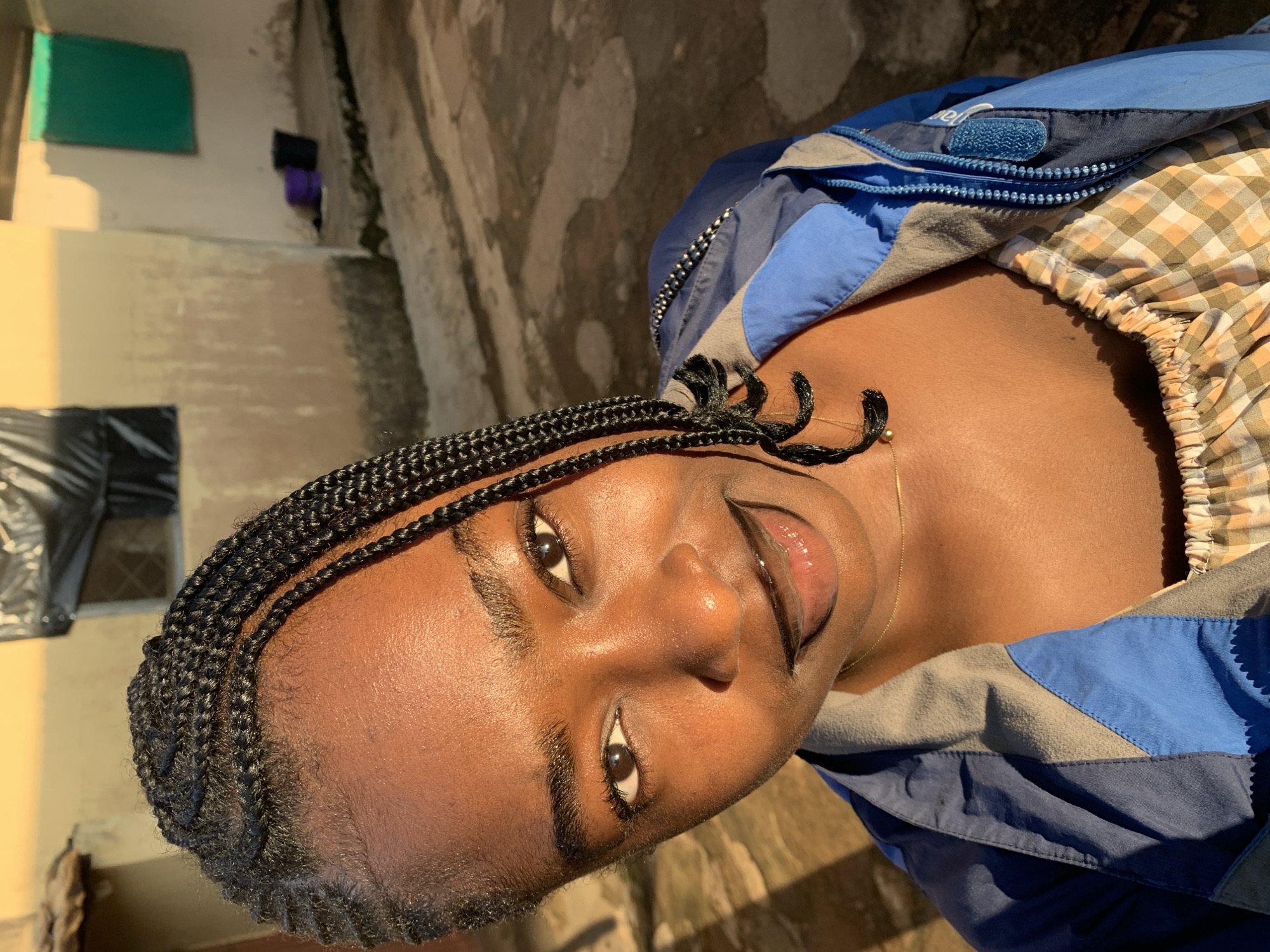 A young woman with dark skin and braided hair is shown from the chest up, angled towards the camera. She is wearing a blue and gray jacket over a checkered top. Her lips are painted with dark lipstick. The background appears to be an outdoor setting in what is described as a city in Rafin Par, Nigeria. The environment is somewhat unkempt, with a concrete ground and building walls visible. Sunlight casts shadows and highlights on the scene.