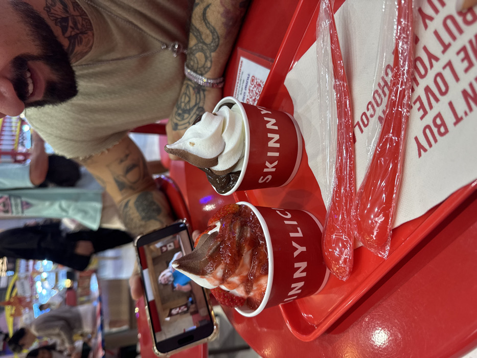 The image depicts a close-up scene inside a Skinnylicious dessert shop, located within a commercial establishment in Bangkok, Thailand. A male individual, visible from the chest up, with a dark beard, extensive tattoos on his right arm and neck, and wearing a light-colored sleeveless shirt, is seated at a glossy red table. His right hand holds a gold-colored smartphone horizontally, displaying a video of a bearded man in a light blue shirt giving a thumbs-up.
On a red plastic tray in front of him are two red cups of soft-serve dessert, both branded "SKINNYLICIOUS" in white text. The left cup contains white and brown swirled soft-serve generously topped with strawberry sauce and fresh strawberry pieces. The right cup contains white and brown swirled soft-serve with a dark chocolate drizzle around its base. Also on the tray are two individually plastic-wrapped red plastic spoons and a white napkin with red text partially reading "DON'T BUY ME LOVE BUT YOU". A small red card containing a QR code is visible next to the right dessert cup. The background is blurred, showing other patrons and the brightly lit interior of a commercial space, consistent with a shopping mall or food court.