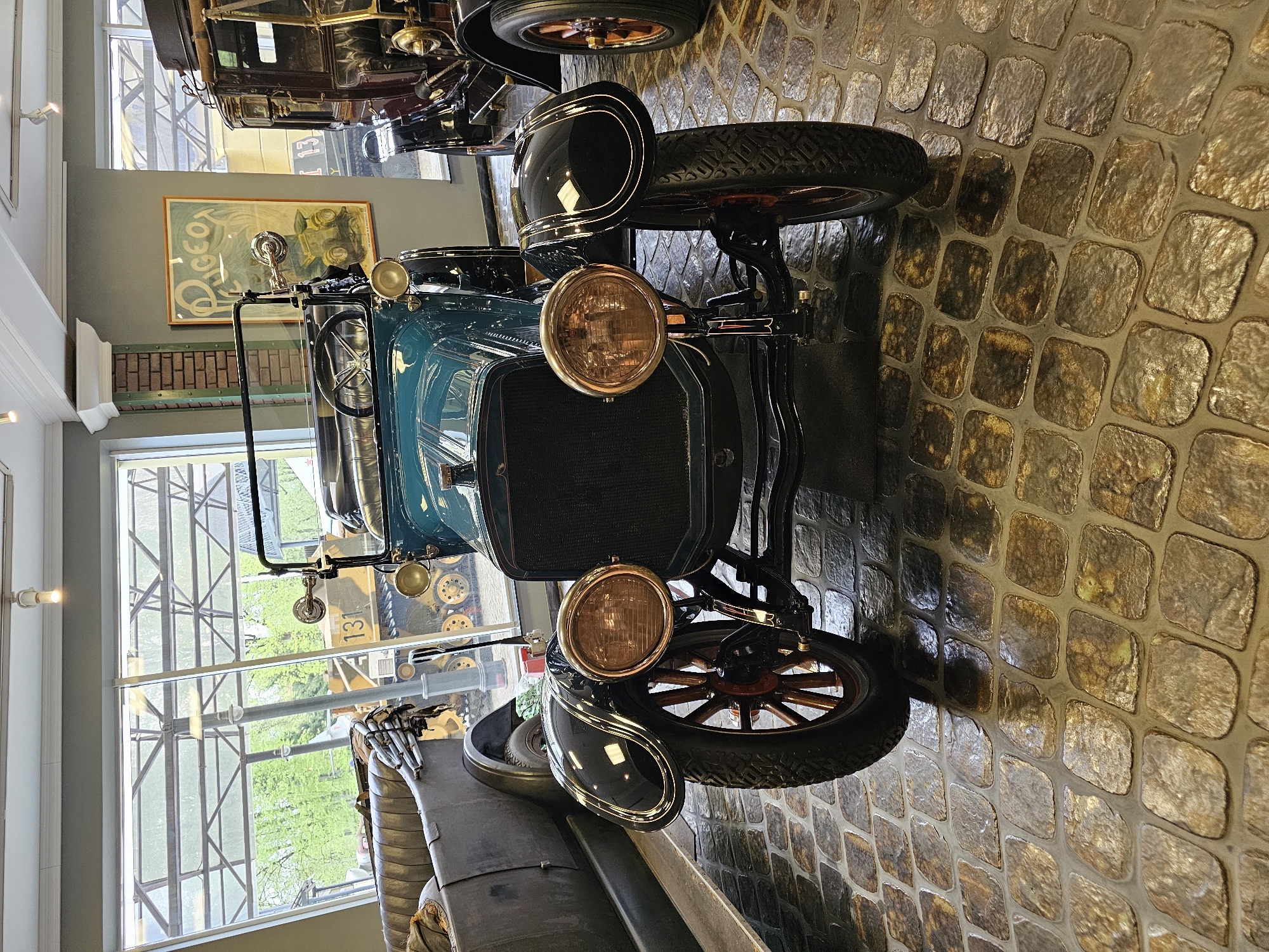 The image showcases a vintage car, possibly a Model T Ford, displayed inside a building, likely a museum, in Архангельское, Russia. The car is teal in color with black fenders and prominent brass headlights. It's positioned on a cobblestone floor. In the background, a large window provides a view of greenery, and another vintage vehicle can be seen through the window, along with a painting on the wall. The overall setting evokes a historical display or exhibit.