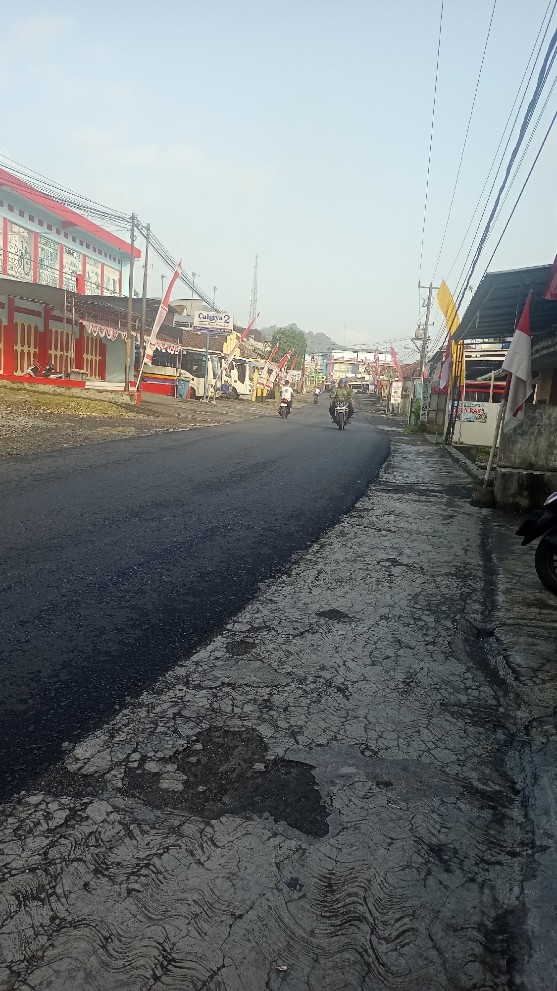 The image depicts a street scene in Karyamandala, Indonesia, under a clear sky. The road in the foreground is paved with asphalt. To the left, a building with a red roof and white walls is visible, featuring red doors and decorative elements. Several red and white flags are displayed along the building and roadside. Further down the street, there are buses and motorcycles. On the right side of the road, a sidewalk area is present, with some parked motorcycles. The overall environment suggests a somewhat developed urban or semi-urban area.