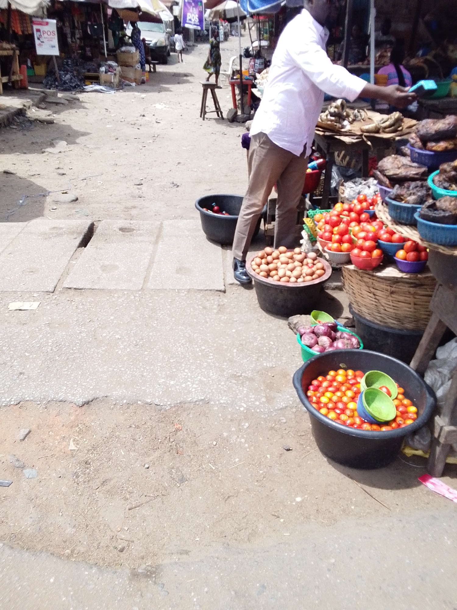 The image depicts a street scene in Lokoja, Nigeria, showcasing a roadside market. A man in a white shirt and brown pants stands behind a display of produce, including tomatoes, eggs, and possibly smoked fish. He appears to be interacting with a customer, holding a blue measuring scoop. The produce is arranged in various black and colorful plastic buckets and baskets. The ground is a mix of concrete and dirt, with some paving stones visible. In the background, there are other market stalls and people, along with signs of commercial activity, including a POS (Point of Sale) advertisement.