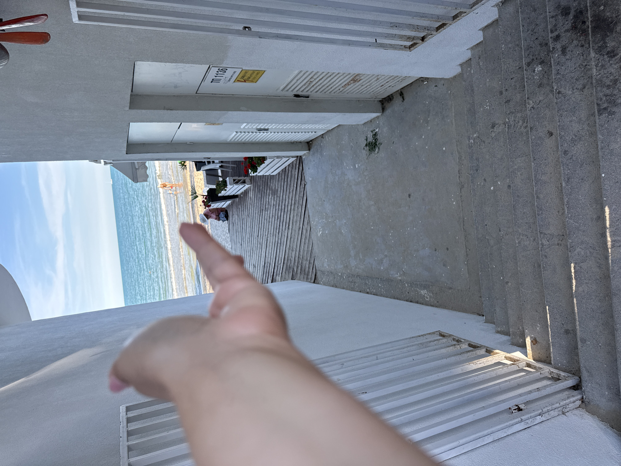 A person's left hand is reaching out into the foreground of the image, obscuring a view of a paved walkway leading down a set of concrete stairs. To the left of the walkway is a white wall with a white metal railing. To the right of the walkway are two white doors with louvers and electrical boxes. A sign with the text "TII 1136" is attached to one of the doors. Beyond the walkway, a wooden deck leads to a beach with a person in a red bathing suit and another person wearing a wide-brimmed hat. The sea is visible in the background, with a calm blue sky above. The scene is set in Varna, Bulgaria.