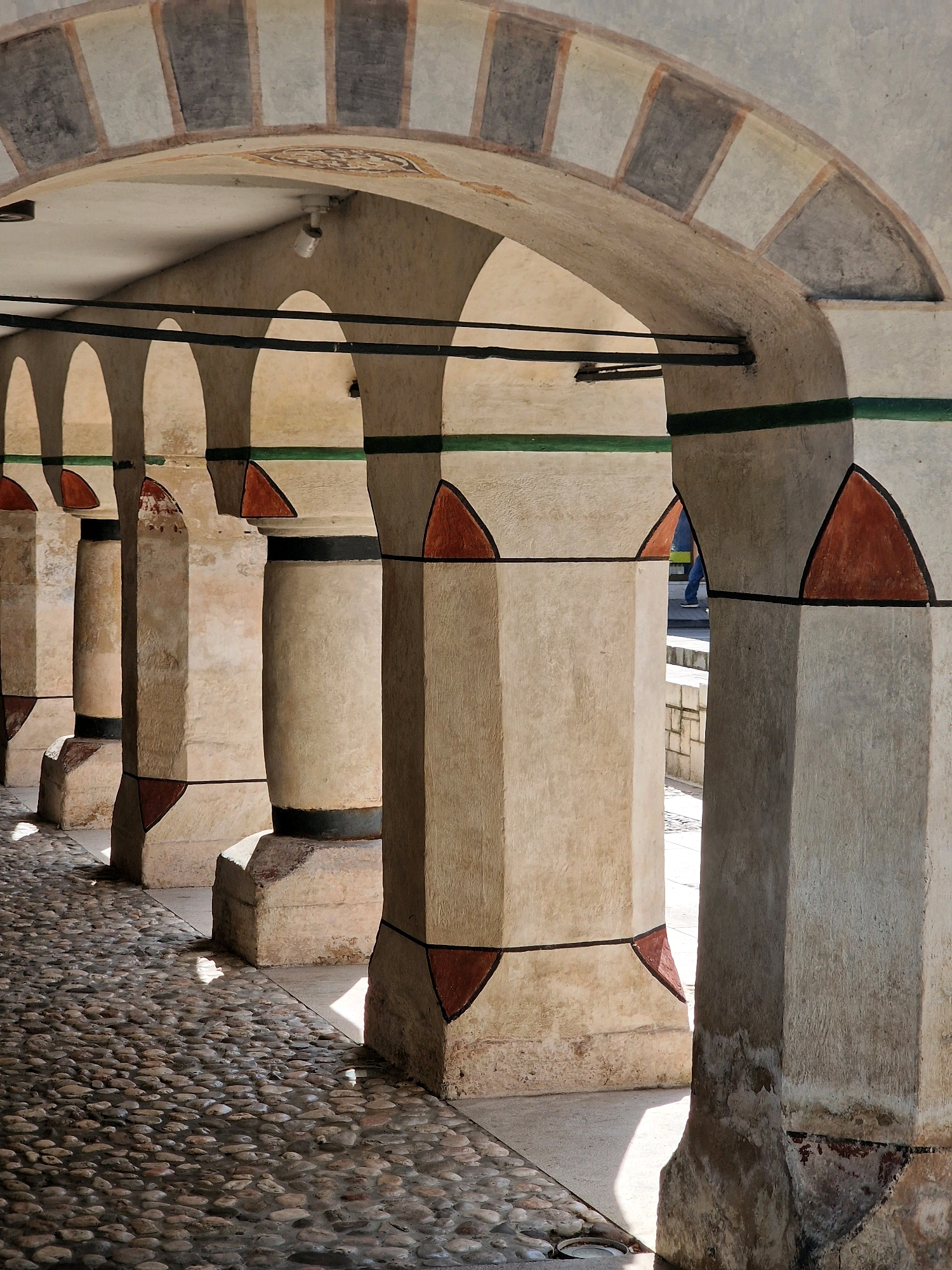 The image shows an arcade with stone pillars and arches, likely part of the Šareni (Multicolor) Mosque in Travnik, Bosnia and Herzegovina. The pillars are square at the base and octagonal above, decorated with triangular terracotta-colored motifs outlined in black. A green band runs horizontally above these motifs, capped by a black band. The arches are decorated with a pattern of alternating light and dark grey squares. The ground is paved with rounded river stones, and sunlight casts shadows through the arcade. In the background, a person in blue jeans and a white t-shirt is partially visible, walking away from the camera. A security camera is mounted on the arched ceiling.