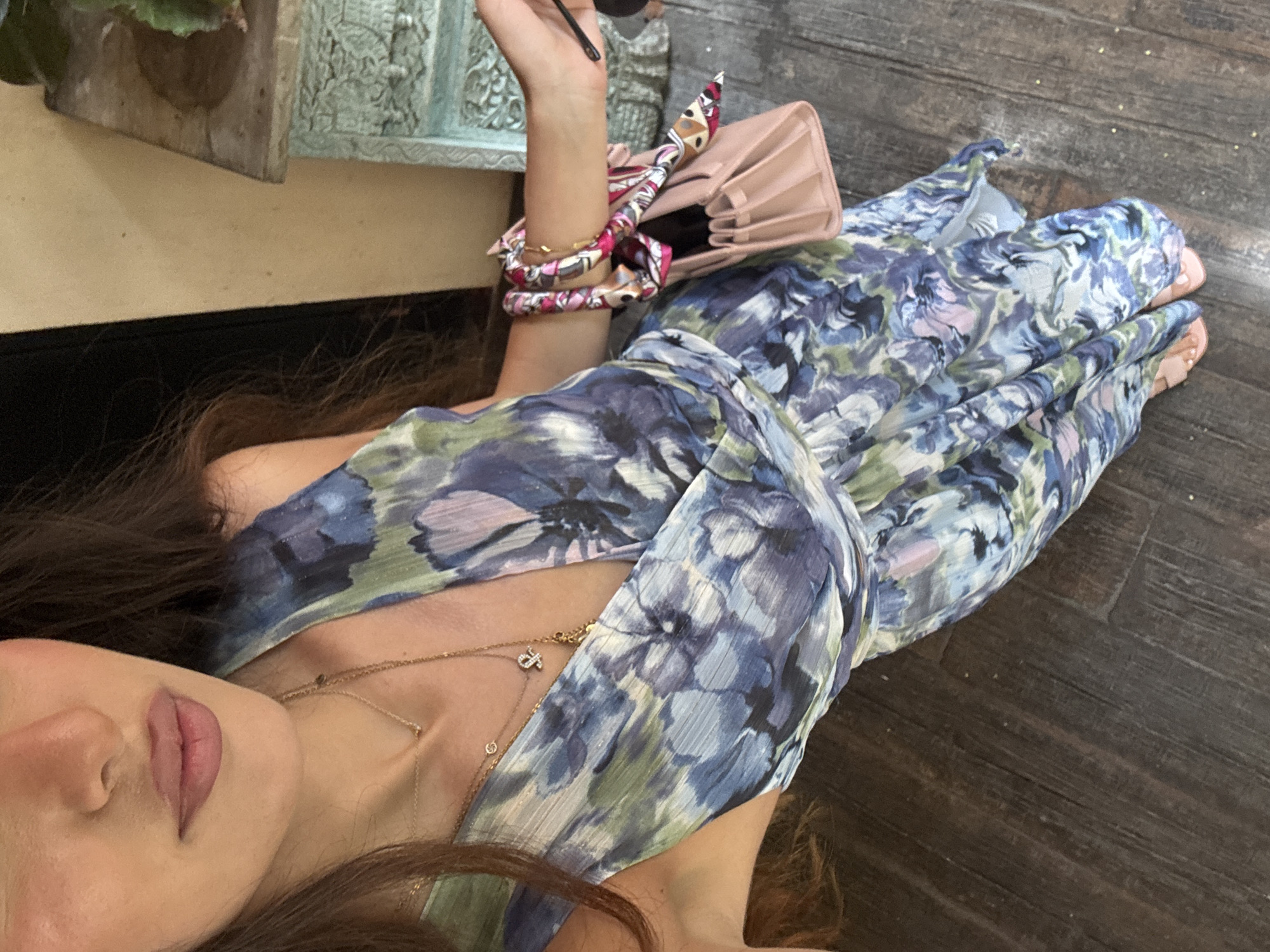 A woman is lying down in a room, seemingly indoors, wearing a light blue and green floral patterned dress. The dress has a deep V-neck, and she is wearing multiple gold necklaces. She has dark hair and fair skin. On her left wrist, she has multiple bracelets and a pink handbag. In the background, there is a wooden floor. The location appears to be indoors in a building in the city of Miami, United States of America.