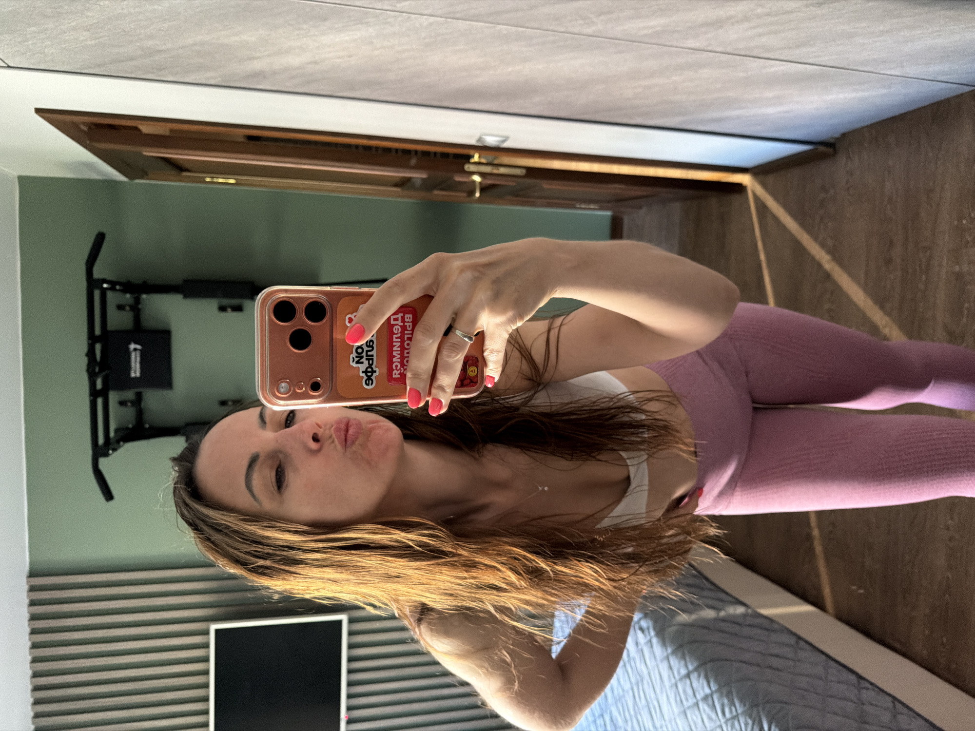 A woman takes a selfie in an indoor environment that may be a residence. She is wearing pink athletic leggings and a white sports top. Her hair is light brown and shoulder-length. She holds a phone in her right hand, with bright pink nail polish on her fingernails. The phone case is decorated with stickers. Behind her, a pull-up bar is mounted on the wall. There is a horizontally striped wall and a television screen. Part of a bed with blue bedding is visible. The floor is light brown wood. A door is visible in the upper background. There is no verifiable evidence in the image to confirm that the location is the city of Birobidzhan, Russia.