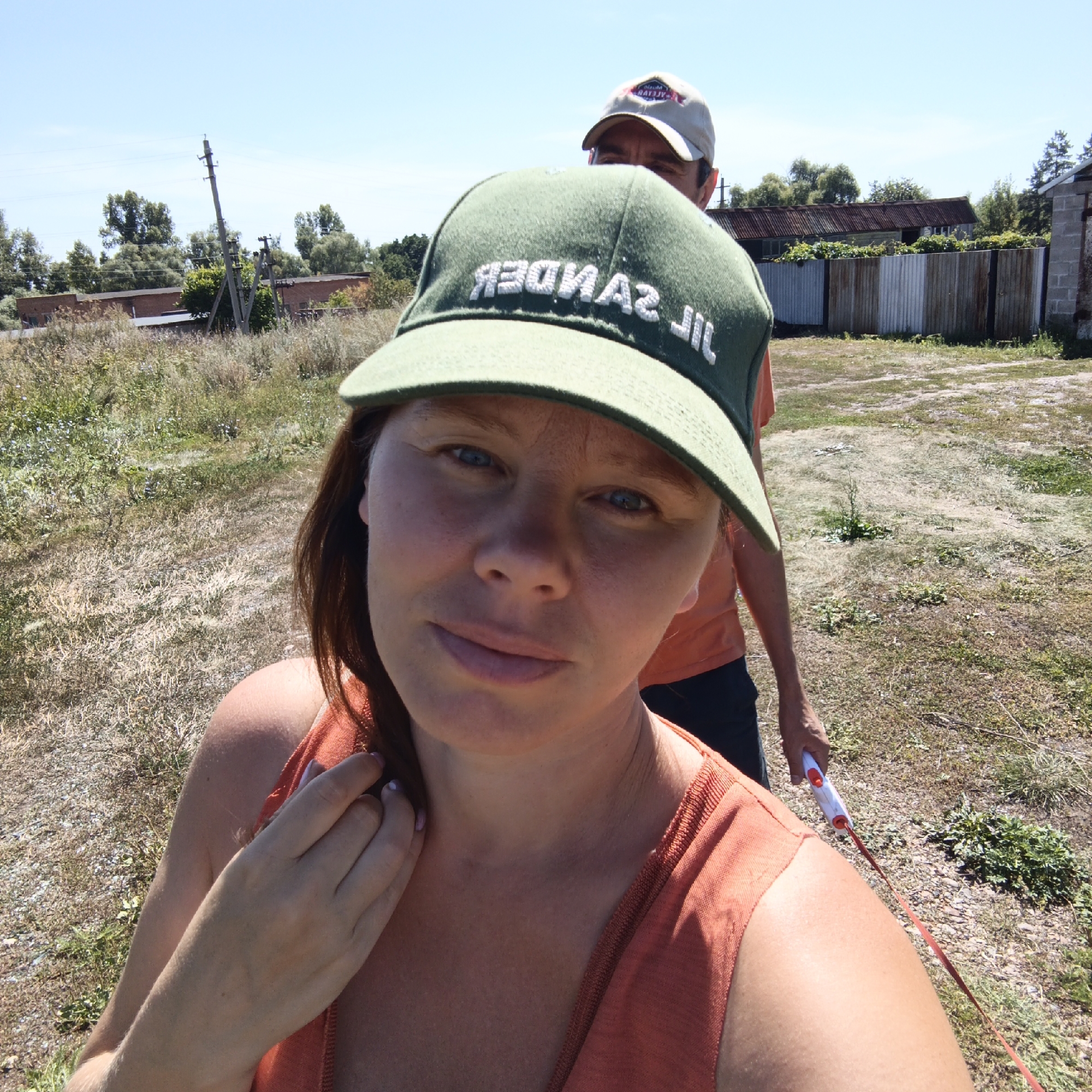 Two people, a woman in the foreground and a man behind her, are outdoors in a rural or semi-rural setting. The woman is wearing an orange tank top and a green baseball cap with white lettering that appears to read "JIL SANDER". She has reddish-brown hair and is looking towards the camera with a neutral expression. Her right hand is near her face, touching her hair. The man behind her is wearing a light-colored baseball cap and an orange t-shirt. He is partially obscured by the woman's cap. The background shows a dry, grassy field with some scattered plants, a corrugated metal fence, and some low-lying buildings. A power pole is visible in the distance. A red retractable leash is being held by the man, extending down towards the bottom right of the frame. The scene appears to be during the daytime under a clear blue sky. The presence of a power pole and corrugated fencing suggests a location in a populated area, potentially a village or the outskirts of a town. Given the context provided, this scene is located in Тугустемир, Russia.