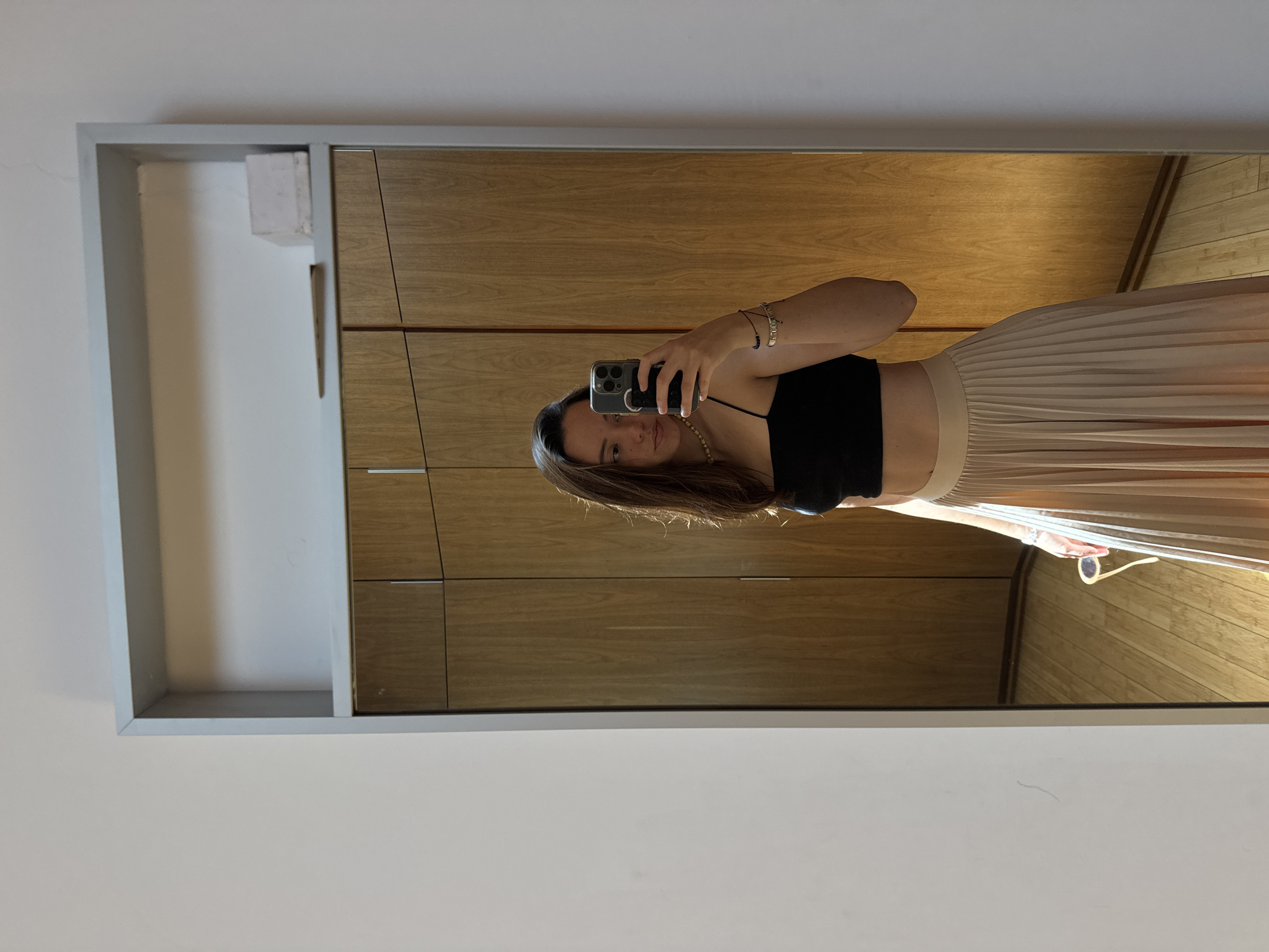 A young woman is depicted taking a mirror selfie indoors in San Bernardino Tlaxcalancingo, Mexico. She stands, holding a modern smartphone with a transparent case in her right hand to capture her reflection. She has long brown hair and is wearing a black crop top, revealing her midriff, paired with a flowing, light-colored pleated skirt, possibly beige or cream. She wears several bracelets on her left wrist and a subtle necklace. Her expression is neutral as she looks towards the phone.
The setting appears to be a well-lit room, likely a bedroom or dressing area, with plain white walls. The woman is standing in front of a large, rectangular mirror framed in light gray. The mirror reflects a wooden wardrobe or closet behind her, which has a warm, light brown finish and segmented panels, some featuring small silver handles. Bright and even lighting suggests it is daytime. A pair of light-colored sunglasses are visible hanging near her right hip, possibly from her skirt or hand. No visible text is present in the image.
