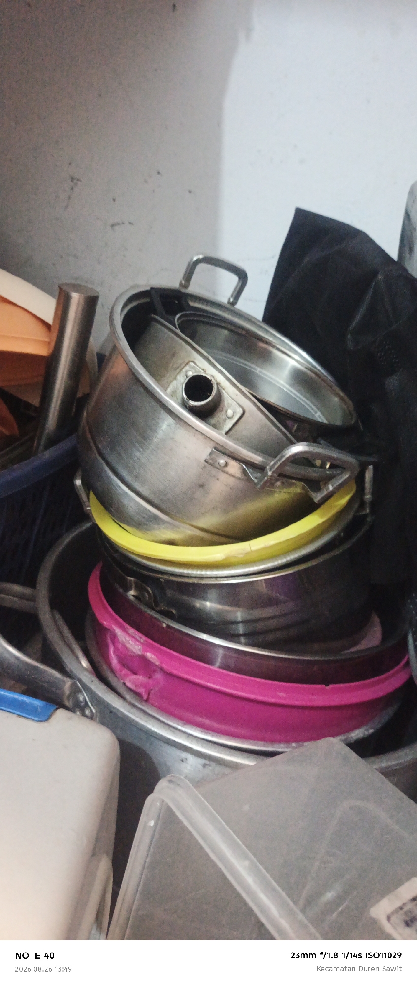 A medium shot captures a stack of cooking pots and plastic containers against a neutral, textured wall. The topmost pot, a metallic silver one with a rounded handle, is tilted, revealing a smaller pot inside. Below it, a bright yellow plastic ring separates it from another metallic pot. Further down the stack, a vibrant pink plastic basin is visible, showing signs of wear. Beneath these, more metallic cookware and translucent plastic containers are stacked. On the left side of the frame, a silver cylindrical object stands upright next to an orange container. The background wall is uneven and appears to be made of concrete. The bottom of the image displays text information, including "NOTE 40", "2026.08.26 13:49", "23mm f/1.8 1/14s ISO11029", and "Kecamatan Duren Sawit". This text suggests the image was taken in the Duren Sawit district, which is part of East Jakarta, Indonesia.