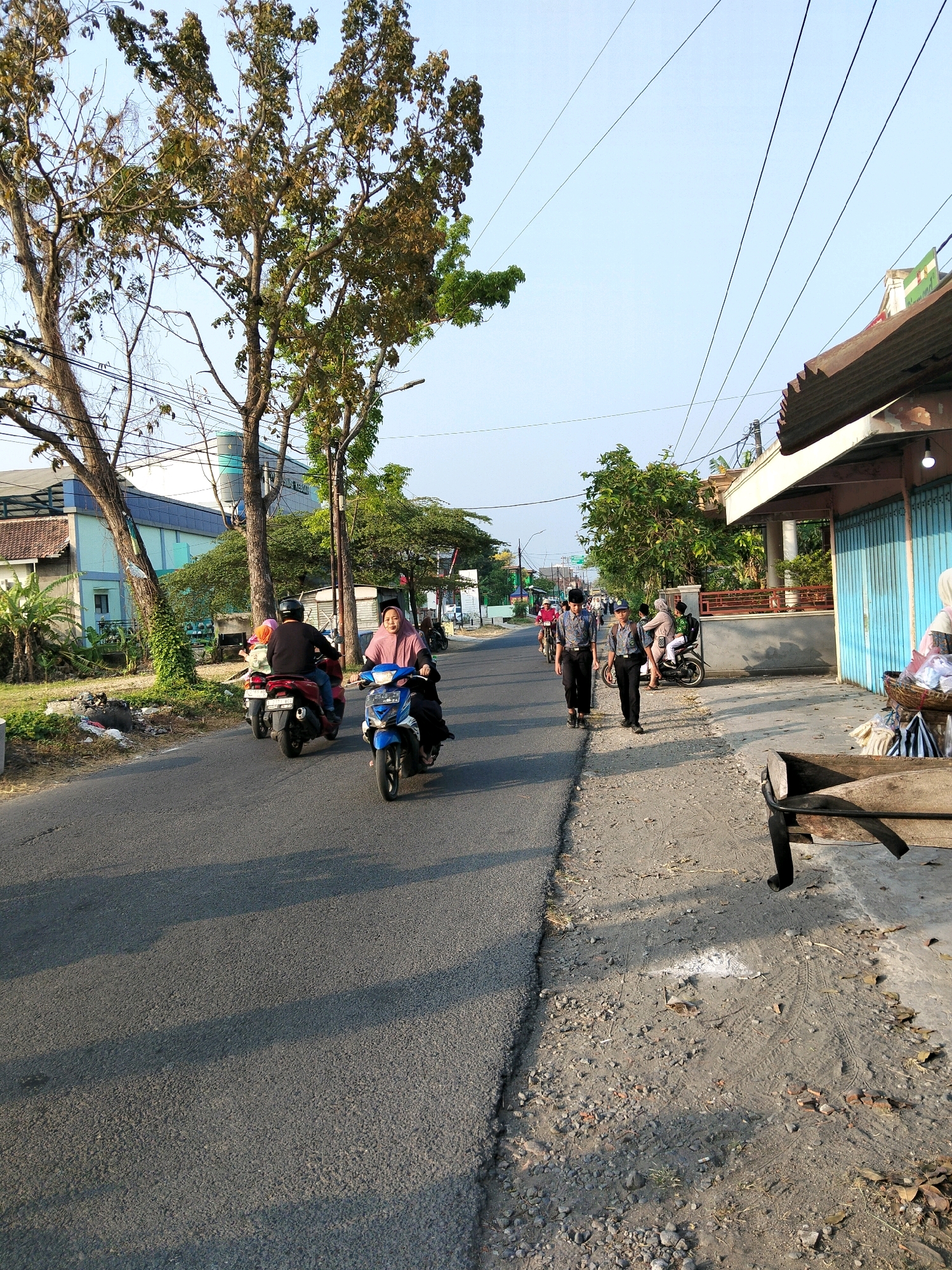 This image depicts a street scene in Keboan, Indonesia, during daylight hours. The sun casts long shadows, suggesting it is either early morning or late afternoon. The road is paved and appears to be a two-lane street with traffic.
On the left side of the road, there is a building with a light blue and white facade, partially obscured by trees and vegetation. A water tower or similar cylindrical structure is visible behind the building. A group of trees with sparse foliage and bare branches line the side of the road, with some greener trees and bushes further back.
Several people are on motorcycles. In the foreground, a woman wearing a pink hijab and a light blue motorcycle is riding towards the viewer. Behind her, another motorcycle carries two passengers, one wearing a dark top and the other a lighter-colored top with a pink headscarf. Further down the road, more motorcycles and a bicycle can be seen, indicating moderate local traffic.
On the right side of the road, there is a sidewalk with a gravel and dirt surface. Along the sidewalk, a building with a corrugated metal roof and light blue folding doors is situated. A wooden bench or table is placed on the sidewalk. A basket filled with items, possibly goods for sale, is also visible near the building.
Two young males in school uniforms, consisting of blue shirts and dark pants, are walking on the sidewalk away from the viewer. Other individuals are also present on the sidewalk and further down the street. Overhead, power lines crisscross the sky. The overall environment suggests a typical urban or semi-urban setting in Indonesia.