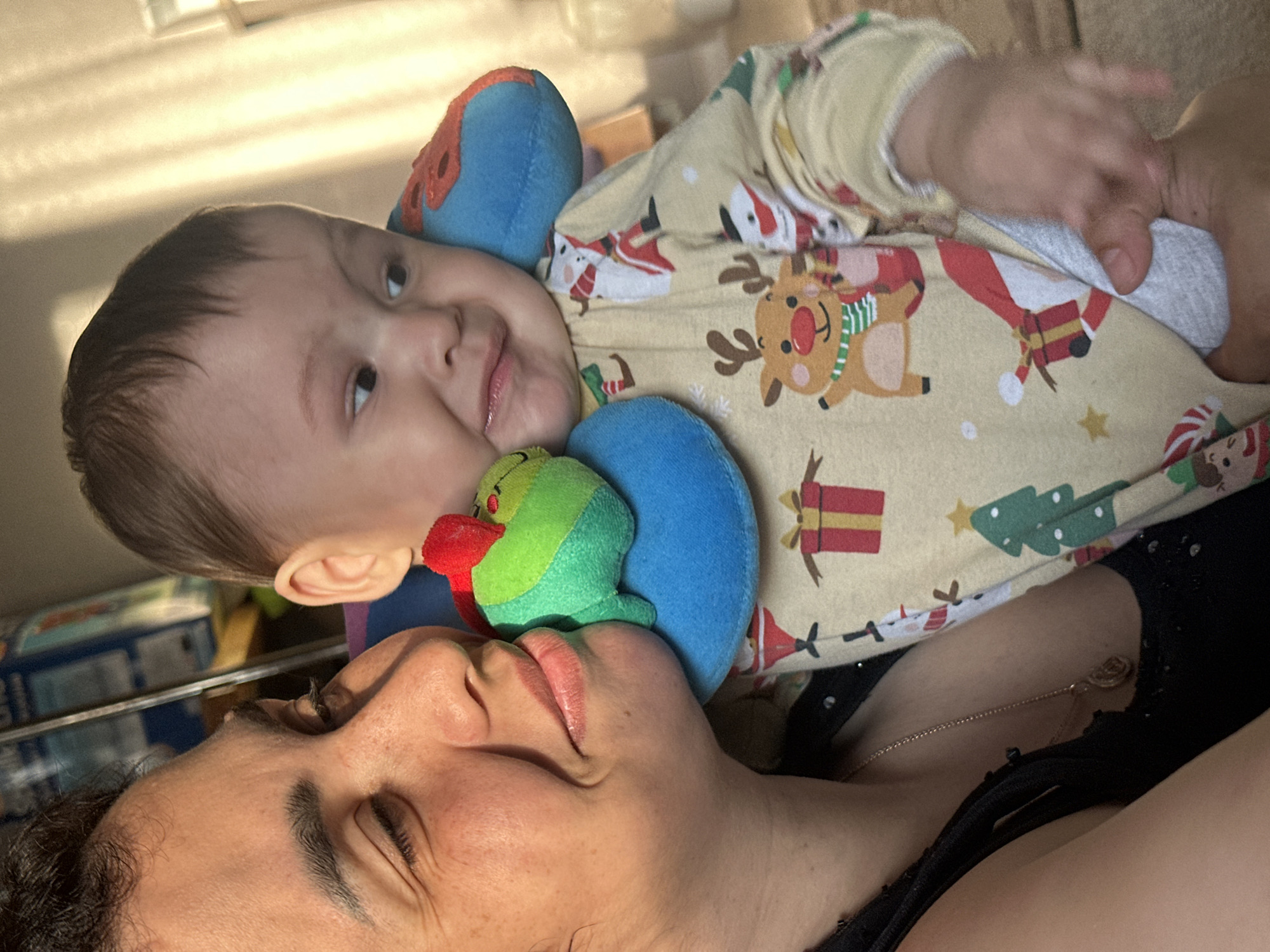 An overhead medium shot captures a woman holding a baby in a room, indoors. The baby is wearing a festive Christmas-themed pajama set with reindeer, snowmen, and gift motifs, and is leaning against a blue travel pillow with a stuffed green creature attached. The woman, positioned on the left, has her eyes closed and a gentle smile, her cheek touching the baby's head. The baby, facing right, exhibits a playful expression, looking away from the camera. The background shows a blurred area with natural light streaming in, creating a warm and soft illumination across the scene.