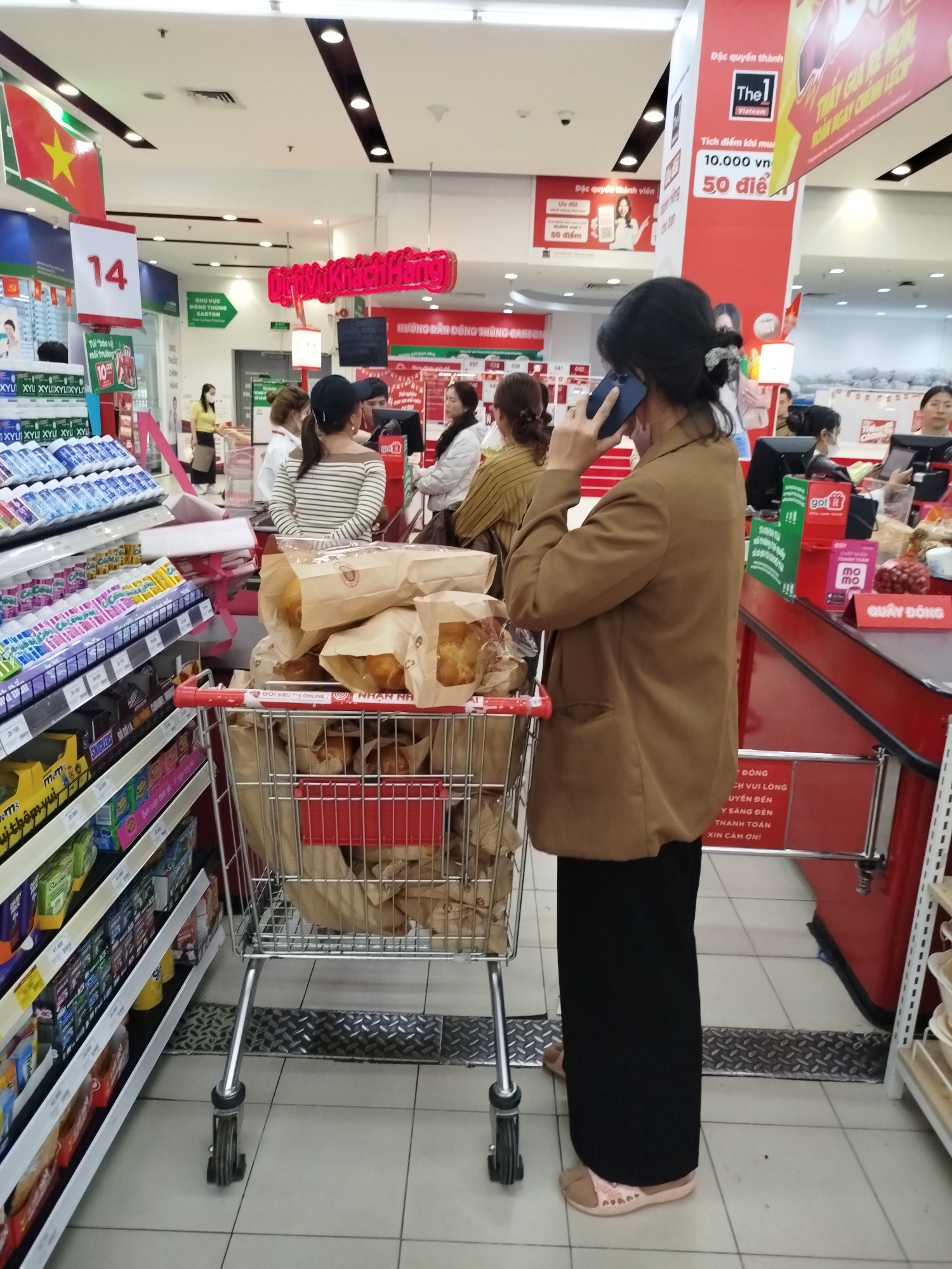 The image depicts an indoor scene inside a brightly lit supermarket in Da Lat, Vietnam, likely at the checkout area. In the foreground, a woman with dark hair tied back, wearing a brown jacket and black pants, stands facing right while talking on a blue mobile phone held to her left ear. She is pushing a metal shopping cart filled with several brown paper bags, which appear to contain baked goods such as bread or pastries. Her foot, wearing a pink slipper-like shoe, is visible near the cart.
In the background, several other customers, mostly women, are visible. Some are queuing at what appears to be a cashier's station, while another woman in a yellow top and face mask stands further back. Supermarket shelves packed with various products like drinks, candies (M&M's, Snickers), and other items line the left side of the aisle.
The setting is characterized by clear signage in Vietnamese. A large red and white sign indicates "14", likely a checkout lane number. Other signs read "DỊCH VỤ KHÁCH HÀNG" (Customer Service) and "KHU VỰC ĐỒNG THÙNG CARTON" (Cardboard box area). A "The1 Vietnam" loyalty program sign advertises earning "50 điểm" (50 points) for every "10.000 vn" (10,000 VND) spent. Payment app logos like "got it" and "mo mo" are seen on the checkout counter. A Vietnamese flag hangs from the ceiling. The overall impression is a typical busy supermarket environment during what appears to be daytime. A sign near the checkout reads "XIN CẢM ƠN!" (Thank you!).