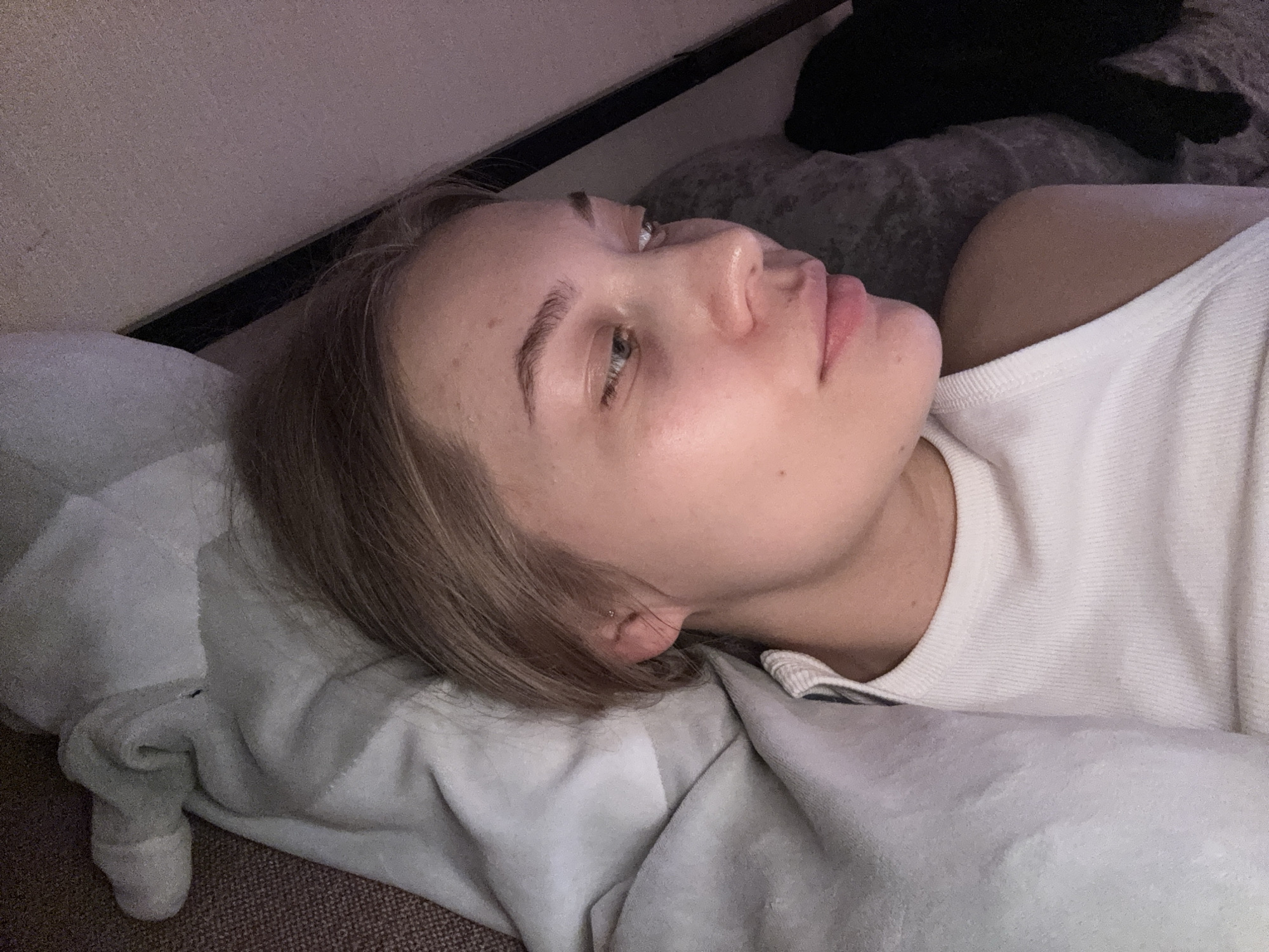 A woman lies on her side on a pillow and blanket. She has light skin, blonde hair, and is wearing a white tank top. Above her on a gray pillow is a black cat. The location of the scene is indoors, but the city of Chaikovsky, Russia cannot be determined from the visual information.