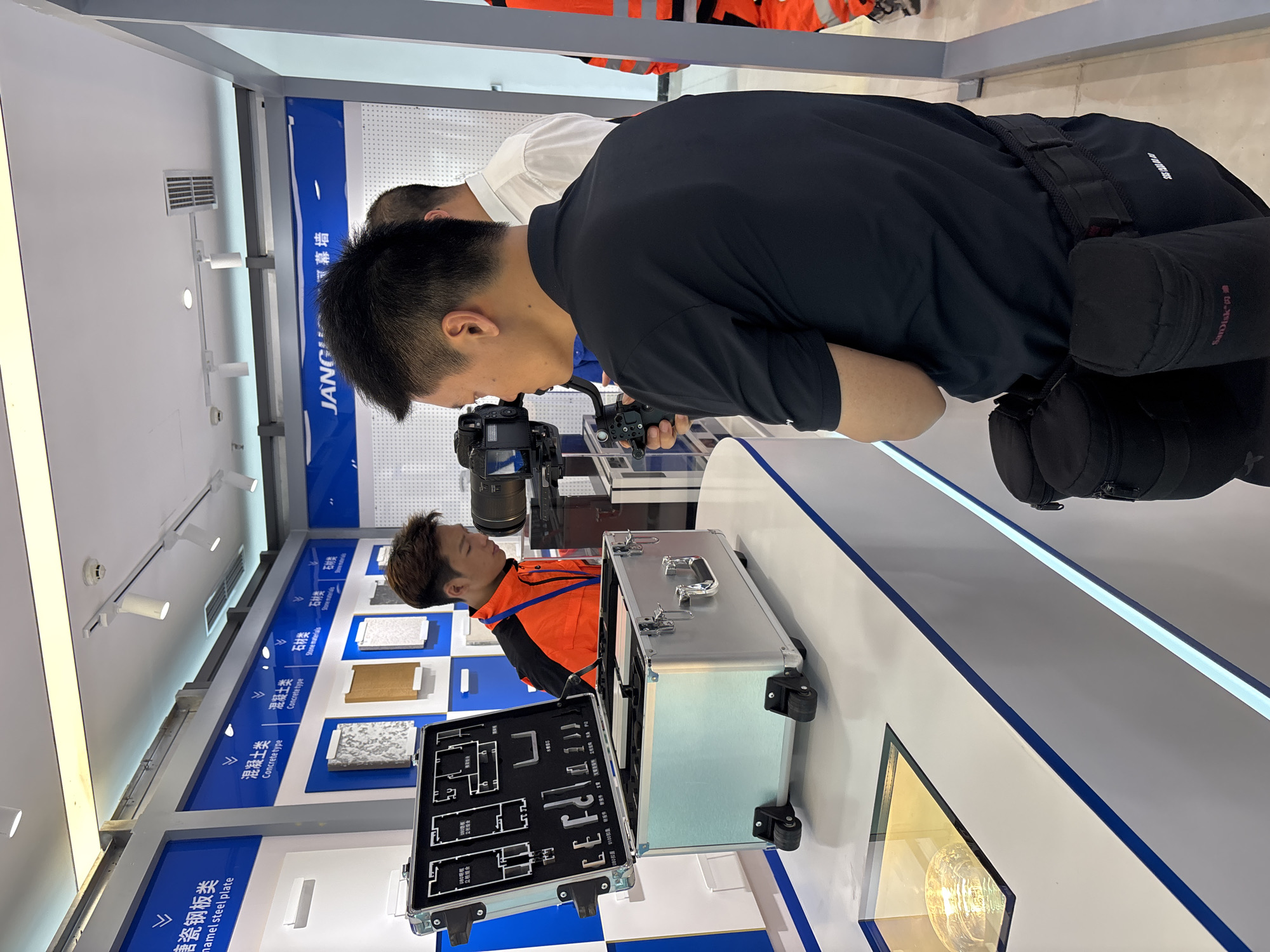 The image shows two Asian men examining a sample case in what appears to be an exhibition or trade show. A man in a black t-shirt is holding a camera with a gimbal and looking closely at the case, which contains architectural samples and diagrams. The case is placed on a display table with blue and white accents.
In the background, there are wall displays with samples of construction materials, labelled in Chinese and English, including "enamel steel plate," "concrete type," and "stone materials."
The environment is brightly lit, and the setting implies an industry-related event or showroom.
There is not enough information in the image to determine that it is in Chengdu, China.