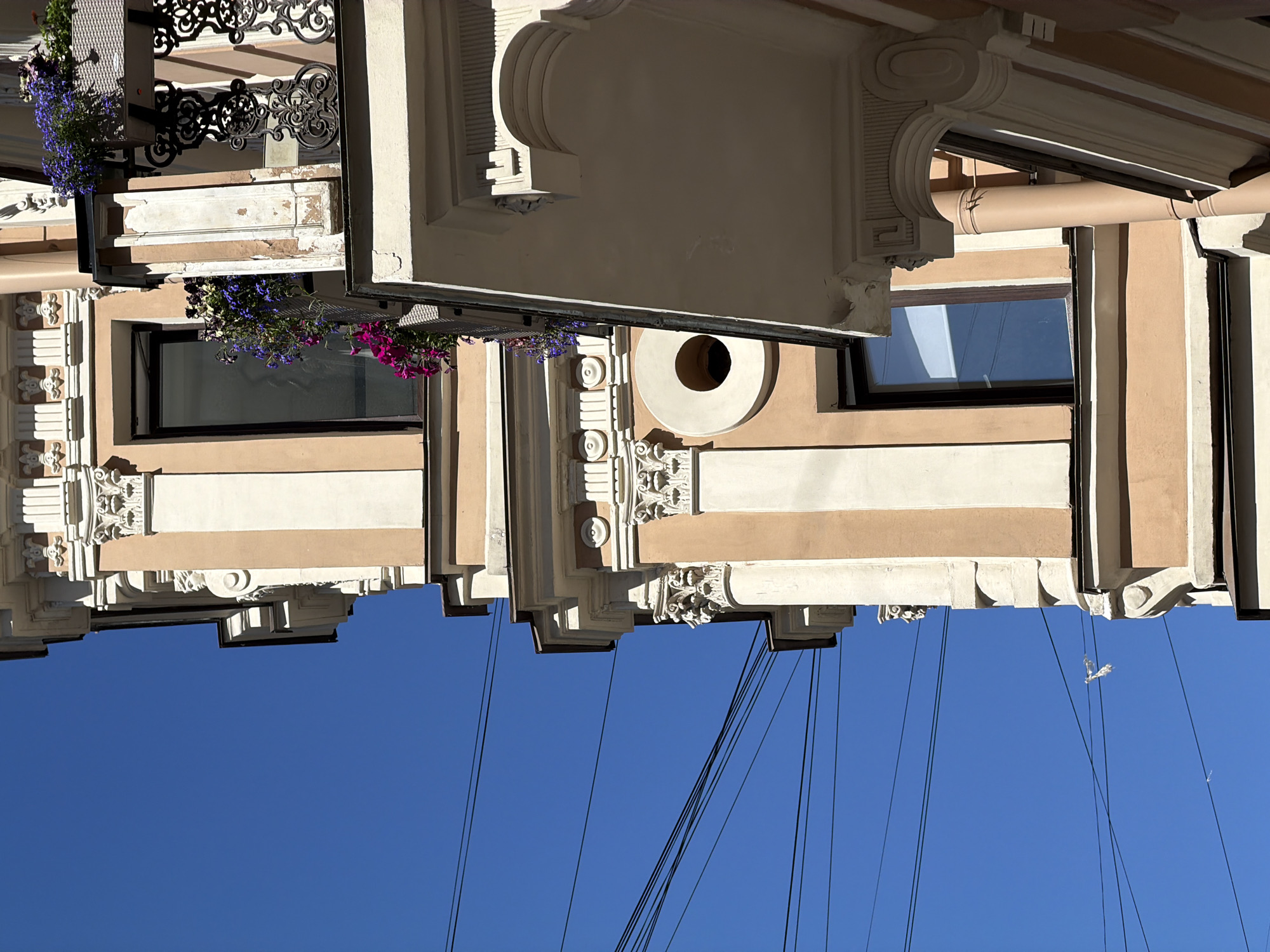 An ornate, multi-story building facade, located at the corner of Rubinstein Street 23 and Grafsky Lane 1 in Saint Petersburg, Russia, is depicted under a clear, deep blue sky. The building exhibits eclectic or Neoclassical architectural elements, with walls painted in alternating vertical sections of light peach and off-white. Numerous decorative details are visible, including pilasters with elaborate capitals featuring acanthus leaves and volutes, decorative friezes, and cornices. Multiple balconies extend from the facade, each supported by decorative corbels, some of which feature sculpted male heads. The balconies are fitted with intricate, dark-colored wrought-iron railings displaying scrollwork and floral motifs. Attached to the balconies and windows are flower boxes brimming with purple, blue, and magenta flowers. A distinctive circular oculus (round window or decorative opening) with a central dark void is prominent on a peach-colored section of the facade. A vertical peach-colored downspout is visible on the lower right. Several black electrical or communication wires traverse the bright blue sky. The scene is illuminated by direct sunlight, casting sharp shadows that accentuate the architectural depth. No people are present.